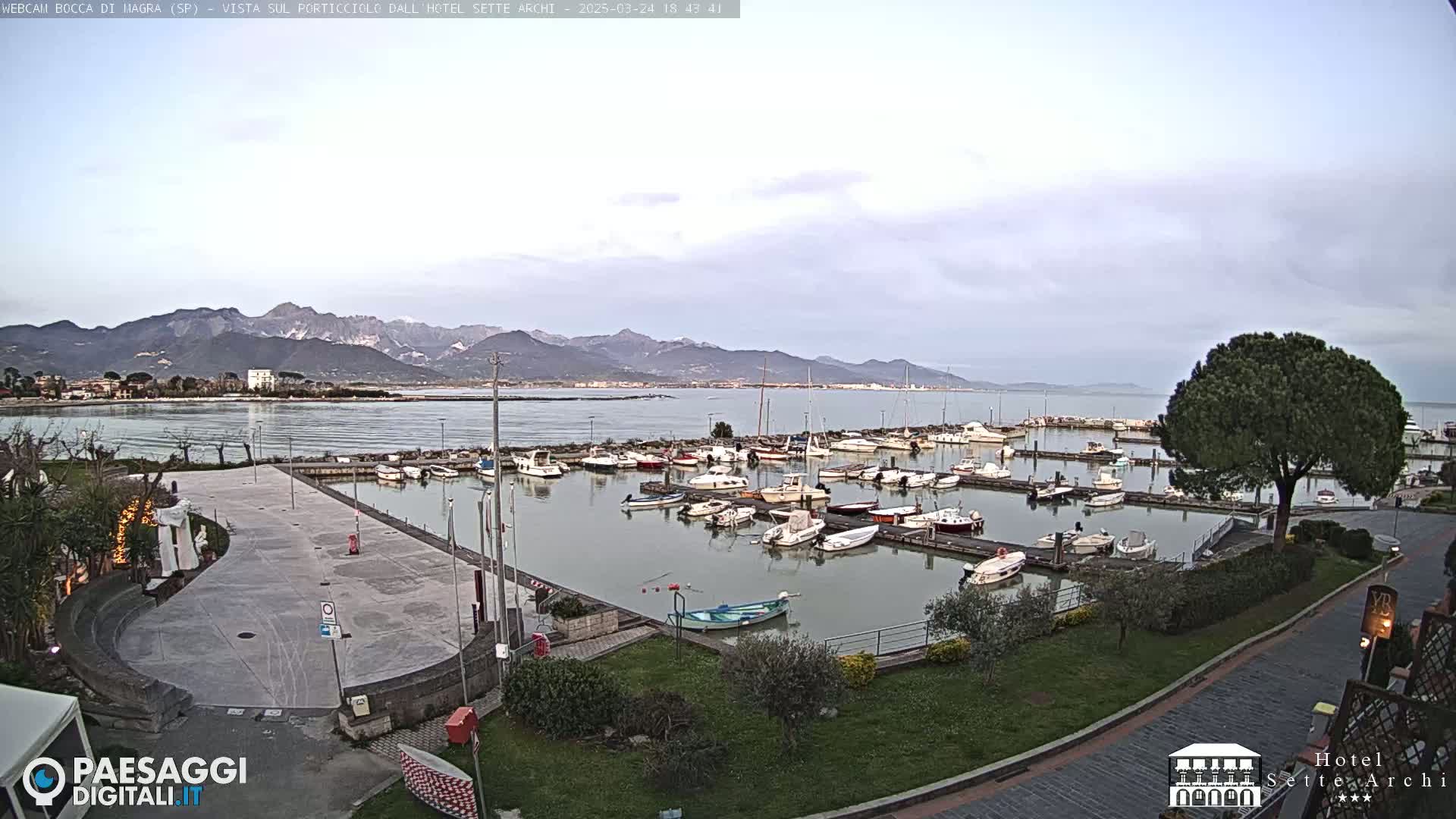 A harbor filled with numerous boats is situated near a paved area, with mountains in the distance under a cloudy sky.
