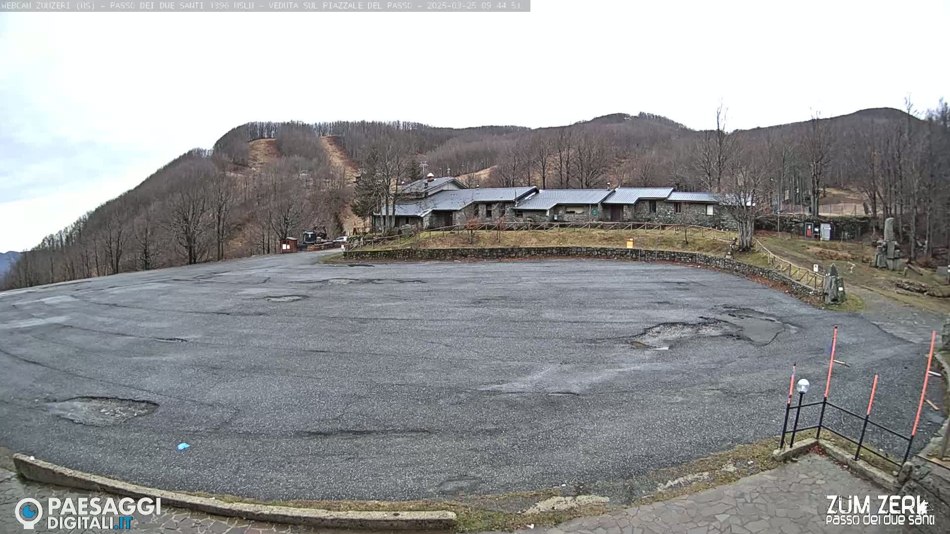 A large, empty asphalt parking lot with potholes sits in front of a stone building, all situated at the base of a treeless, snowless mountain under an overcast sky.