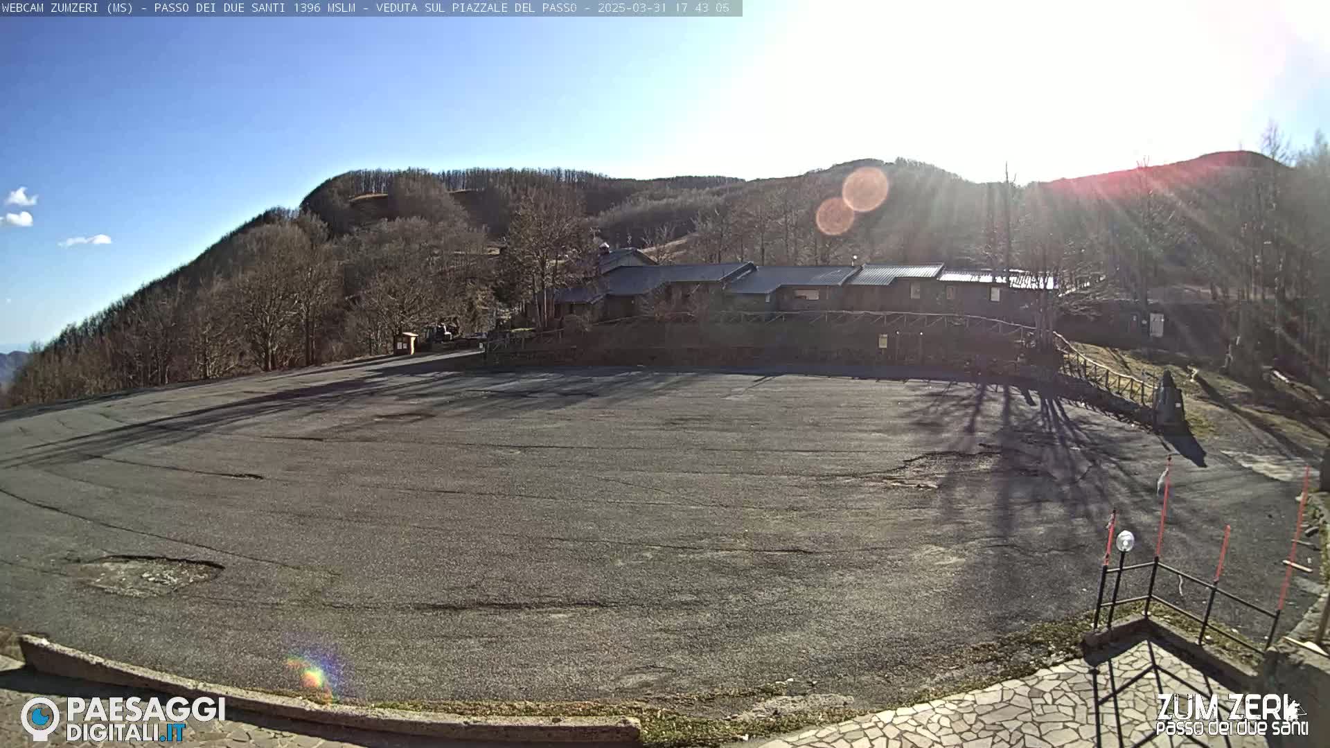 A large, empty asphalt parking lot sits below a cluster of buildings nestled amongst bare, winter trees on a sunny hillside.
