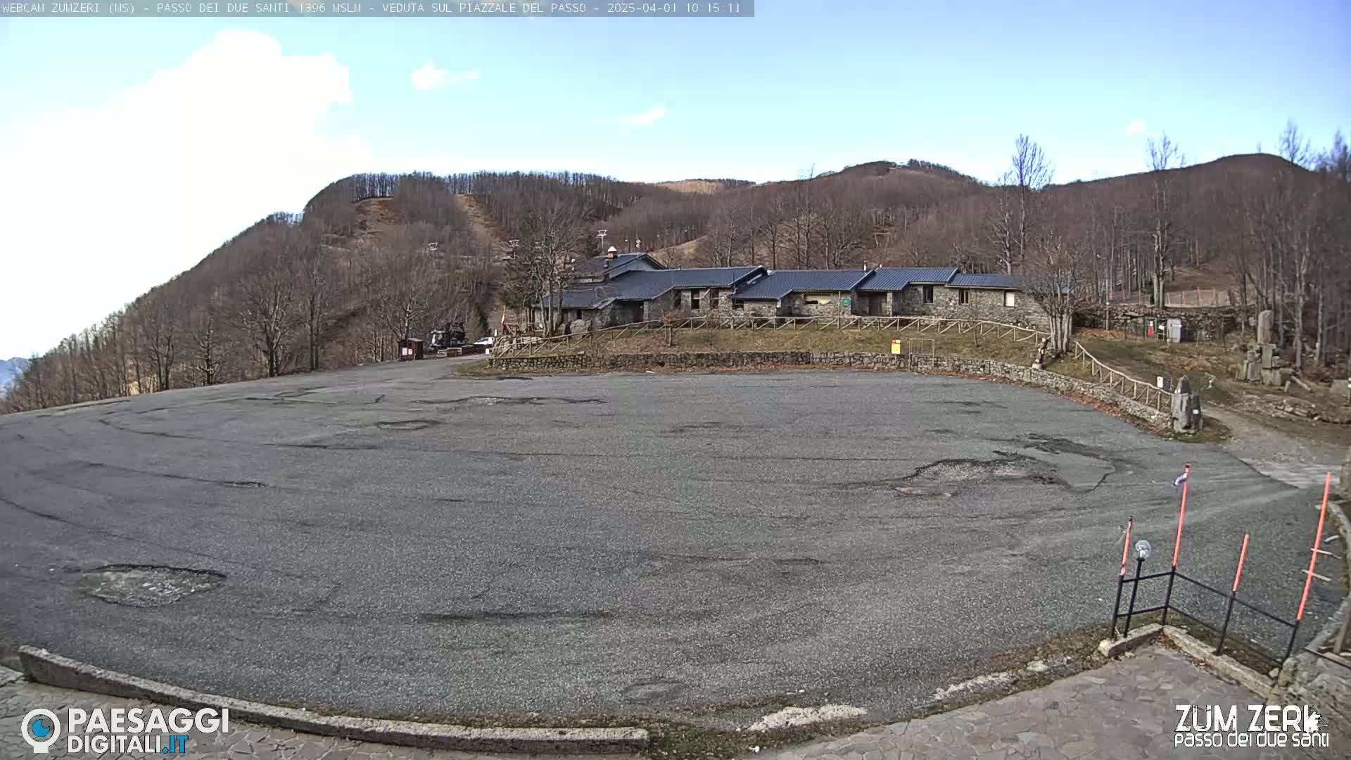 A large, empty asphalt parking lot sits in front of a stone building complex nestled at the base of a sparsely wooded hill under a mostly clear sky.
