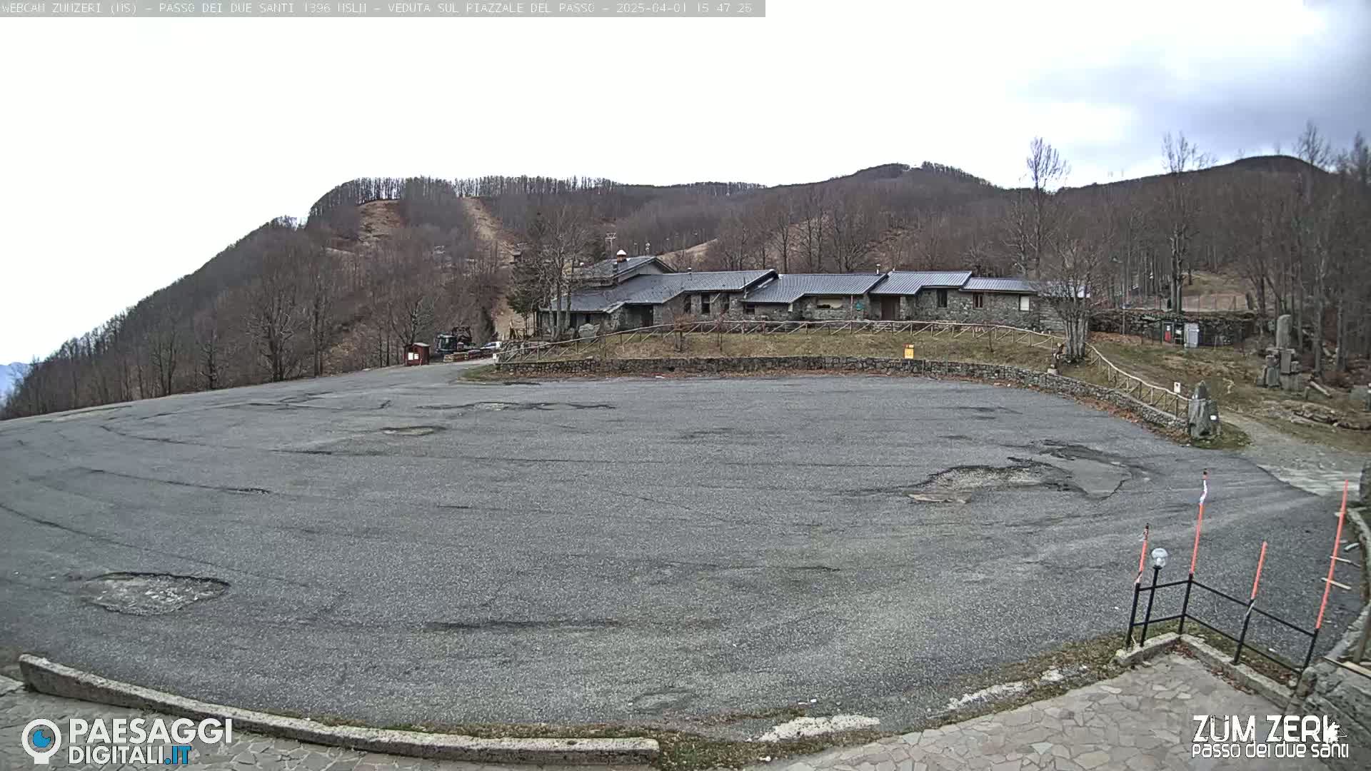A large, empty asphalt parking lot sits before a stone building nestled against a hillside under a mostly cloudy sky.
