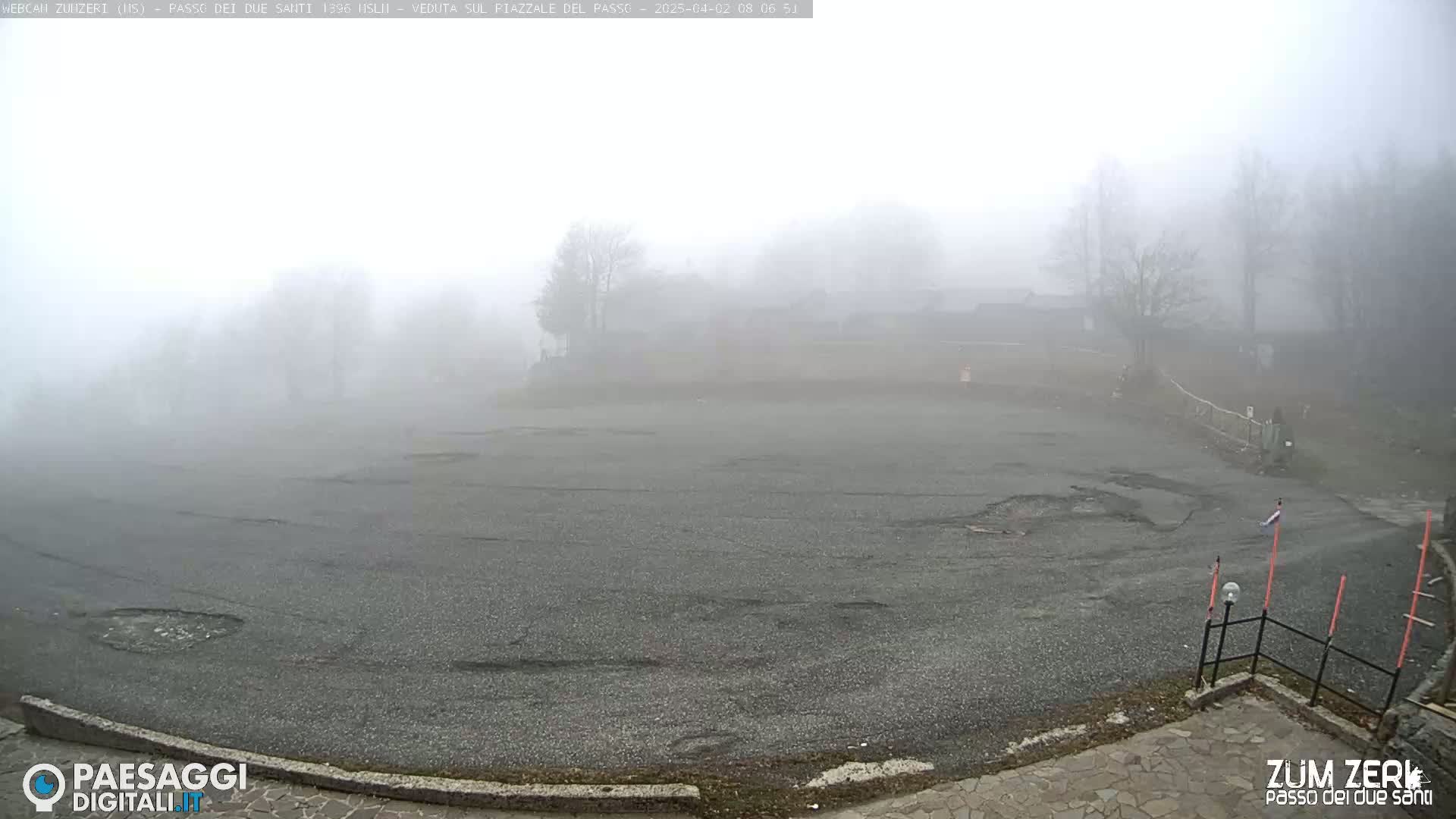 A large, empty asphalt lot is shrouded in thick fog, with hazy silhouettes of trees and buildings visible in the background.