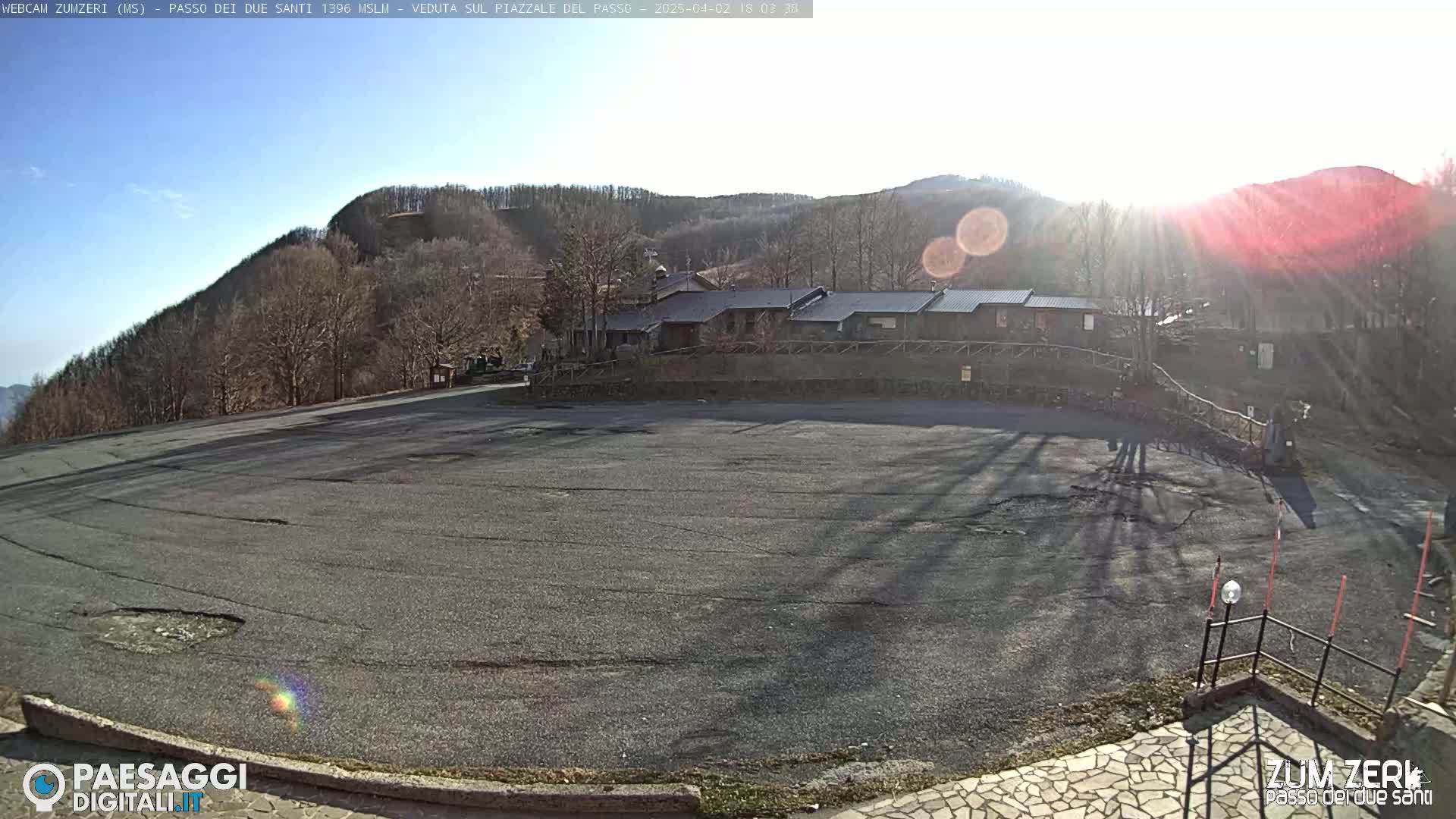 A mostly empty asphalt parking lot sits below a row of buildings nestled in a sunny, mountainous area with bare winter trees.