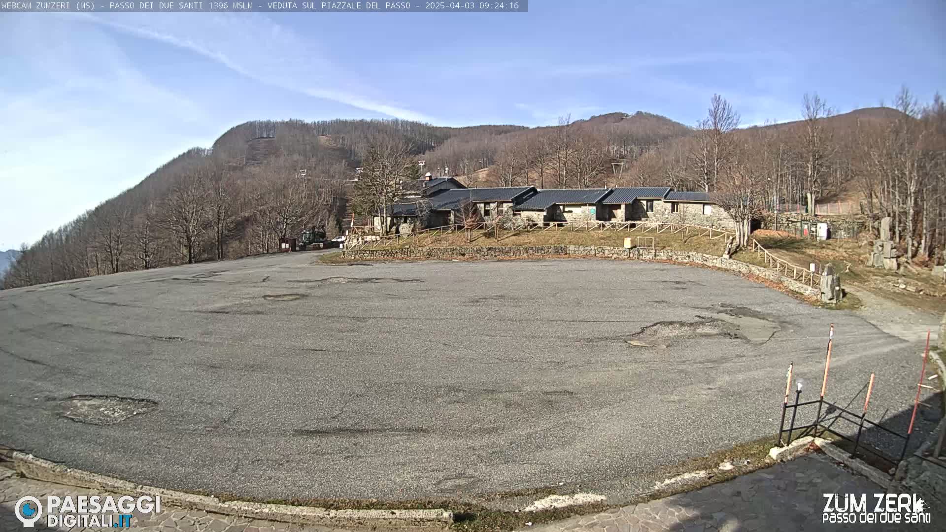 A large, empty asphalt parking lot sits before a stone building complex nestled at the base of a treeless, brown hill under a clear, sunny sky.