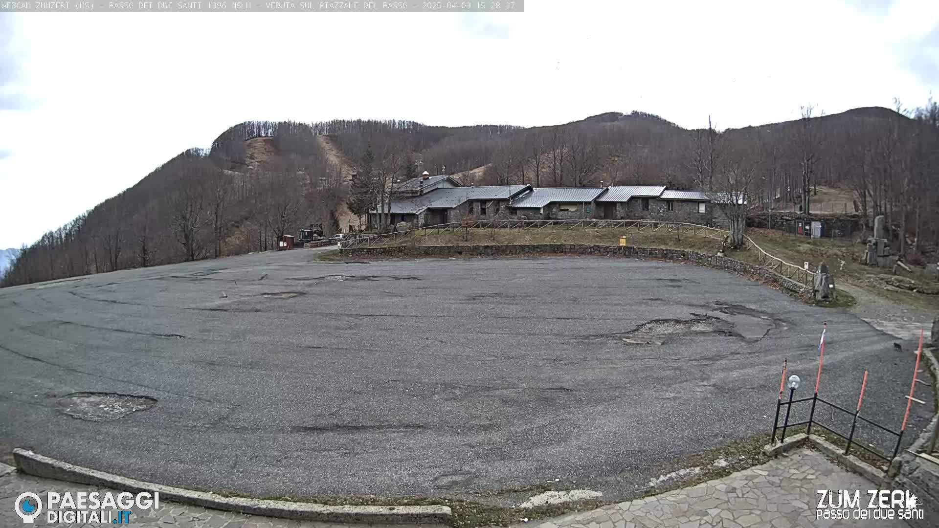A large, empty asphalt parking lot sits before a stone building nestled at the base of a treeless, snowless hill under an overcast sky.