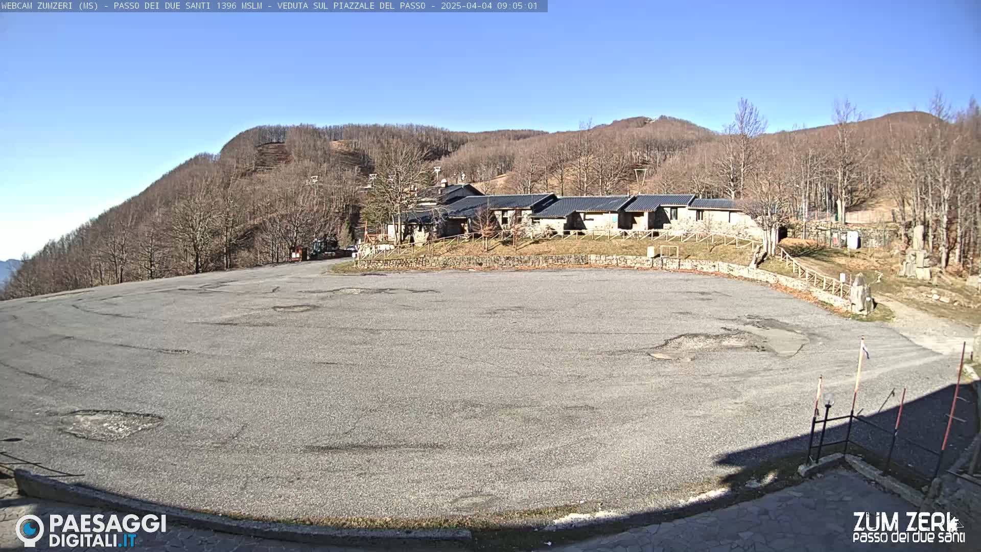 A large, empty asphalt parking lot sits before a cluster of low buildings nestled against a hillside covered in bare, winter trees under a clear blue sky.