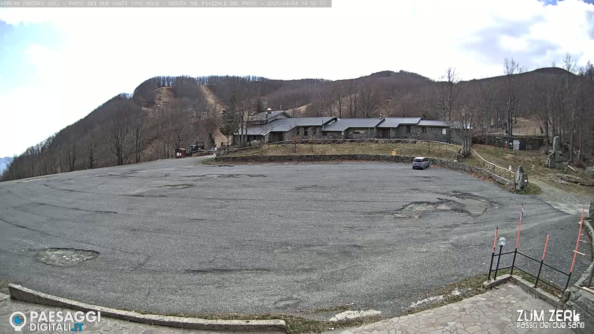 A mostly empty asphalt parking lot sits in front of a stone building complex at the base of a sparsely wooded hill under a partly cloudy sky.