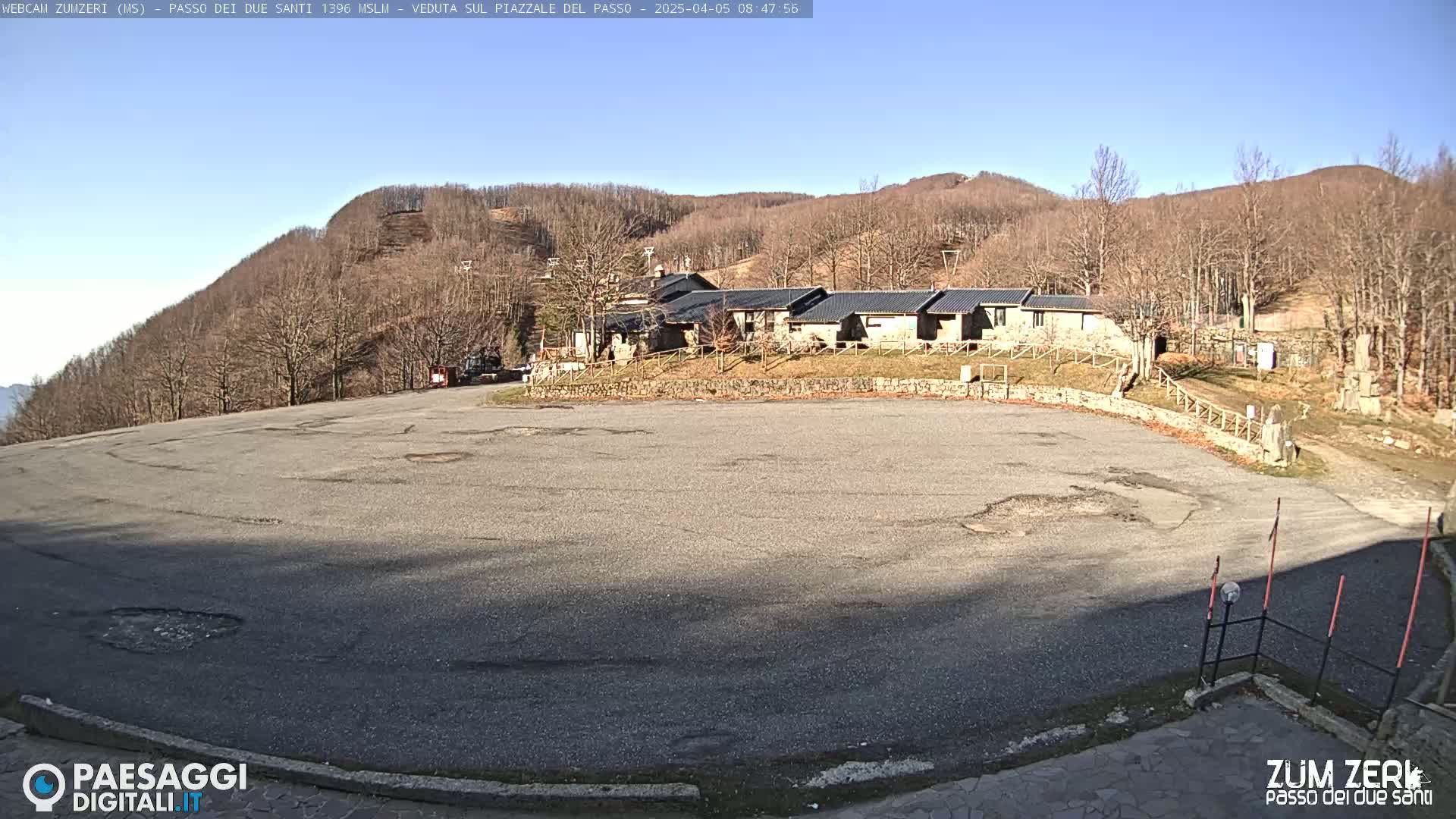 A large, empty asphalt parking lot sits before a cluster of buildings nestled at the base of a hill covered in bare, brown trees under a clear, sunny sky.