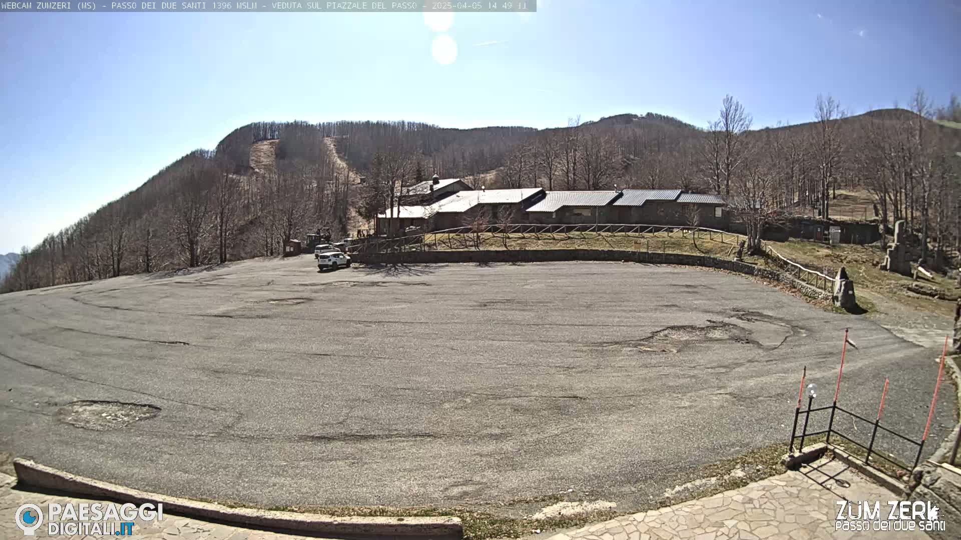 A large, mostly empty asphalt parking lot sits before a long, low building nestled among bare, brown trees on a sunny hillside.