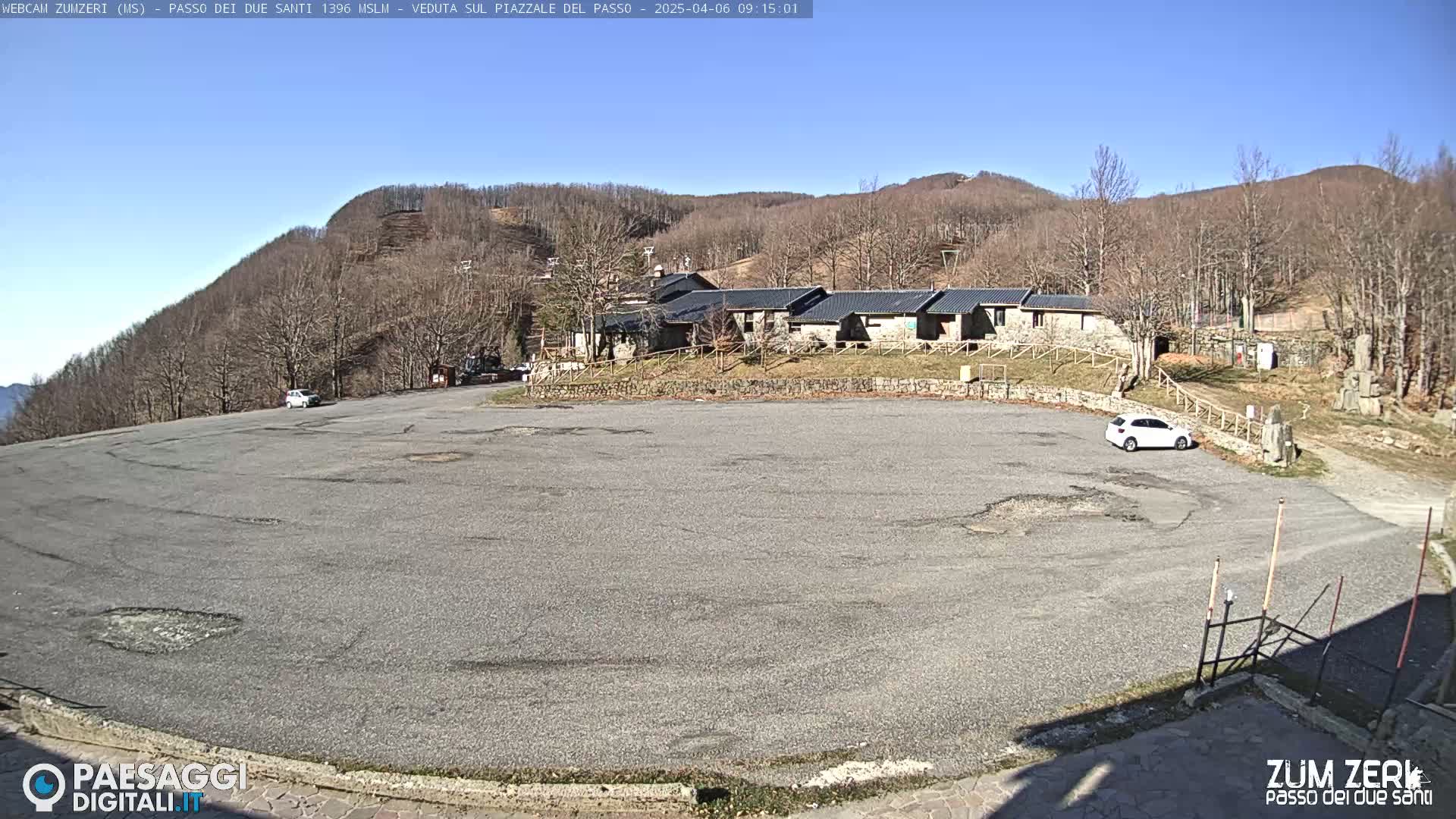 A mostly empty asphalt parking lot sits before a cluster of low buildings, all nestled against a hillside of leafless trees under a clear blue sky.