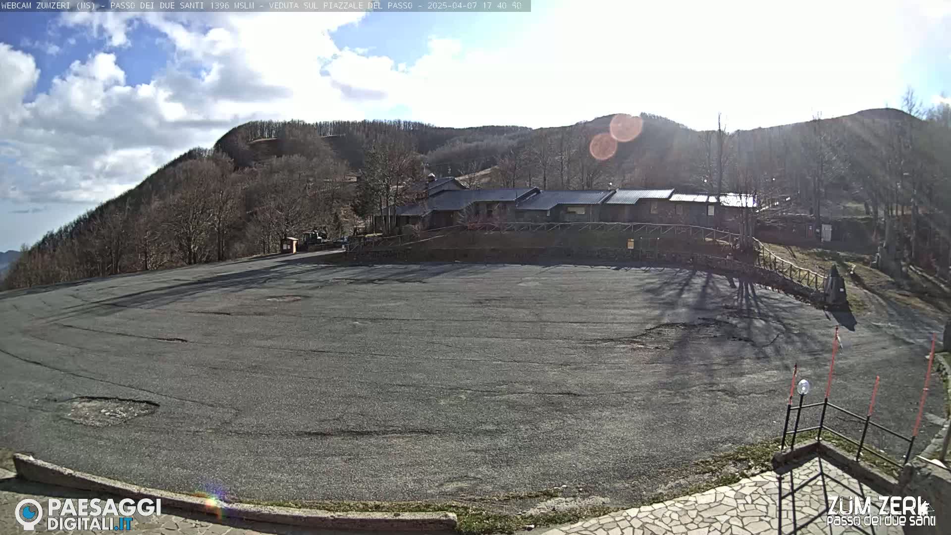 A large, empty asphalt parking lot sits in front of a low building complex nestled against a hillside covered in bare, winter trees under a partly sunny sky.