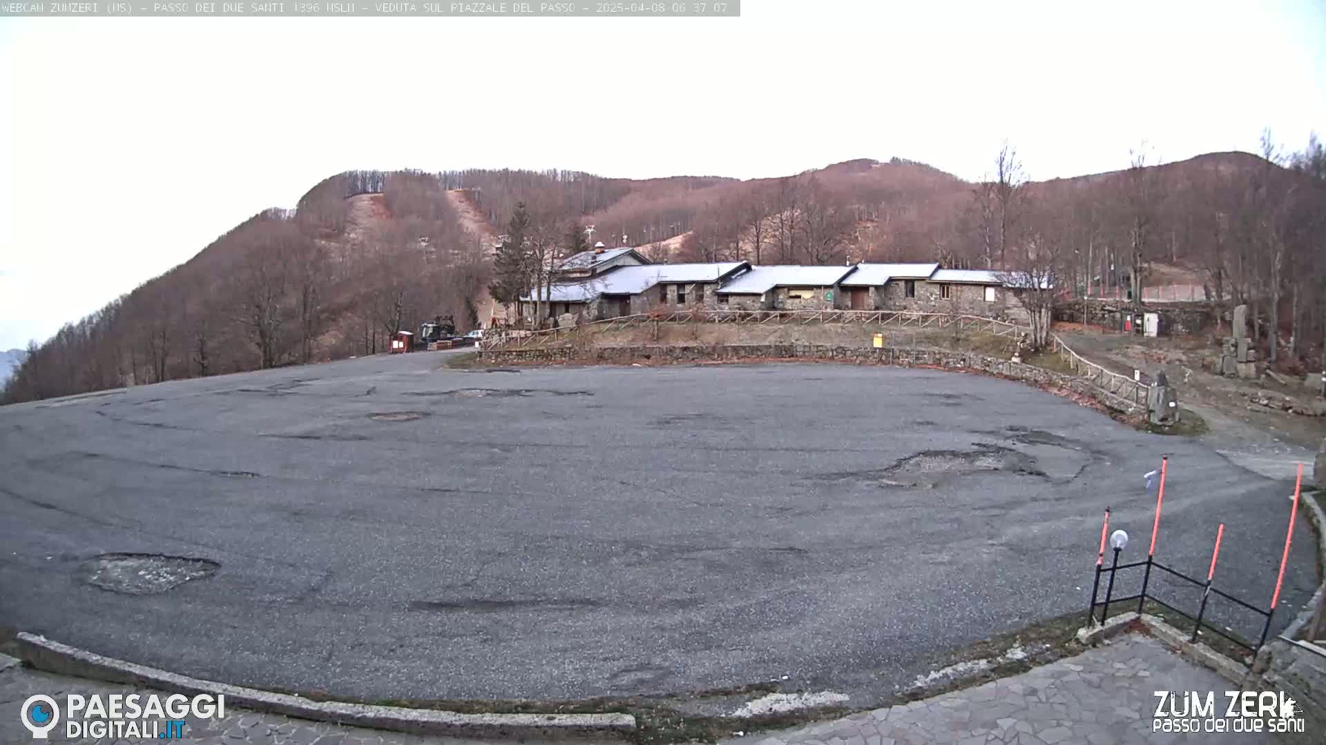 A large, empty asphalt parking lot with potholes is situated in front of a long, low stone building, backed by a treeless hillside under an overcast sky.