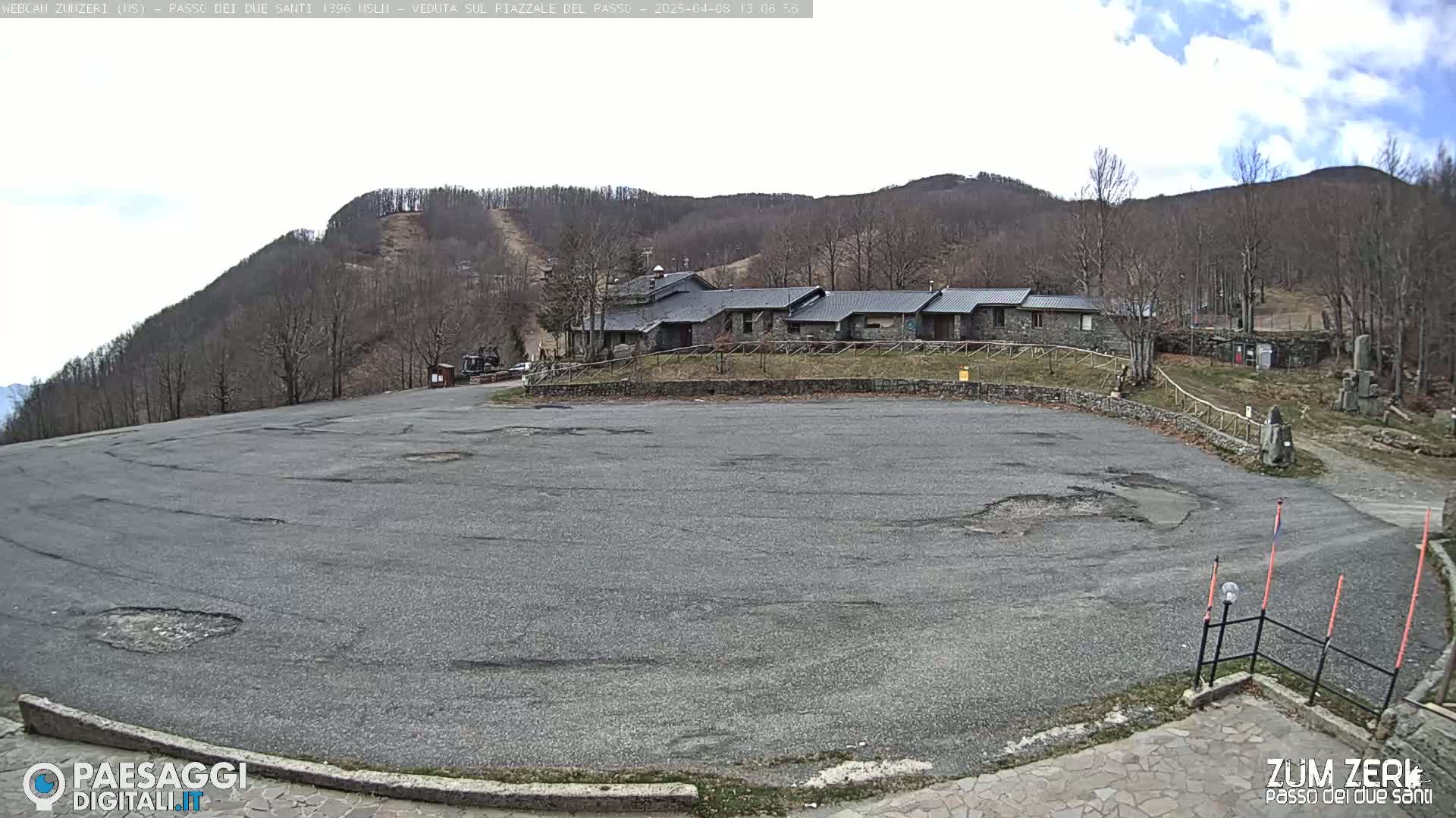 A large, empty asphalt parking lot sits before a stone building complex nestled against a sparsely wooded hillside under a partly cloudy sky.