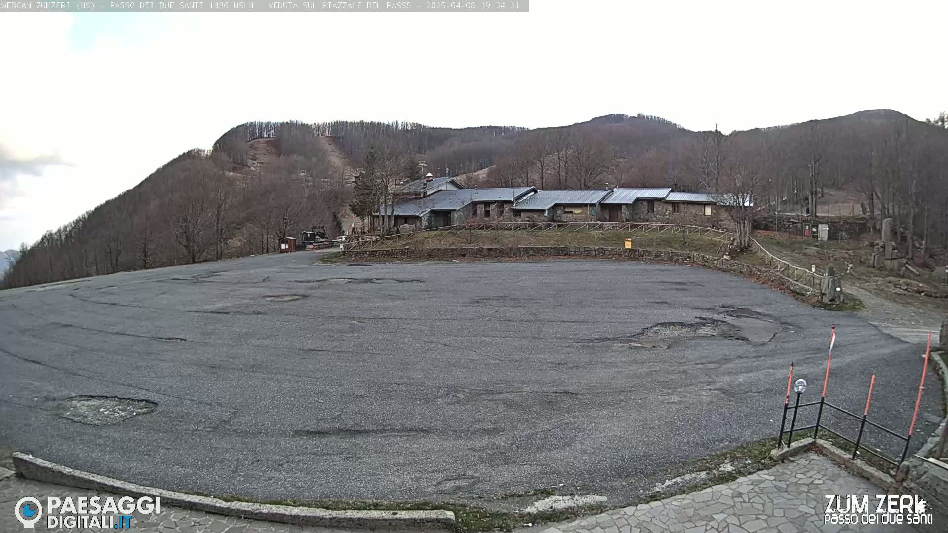 A large, empty asphalt parking lot sits before a cluster of stone buildings nestled against a sparsely wooded hillside under an overcast sky.