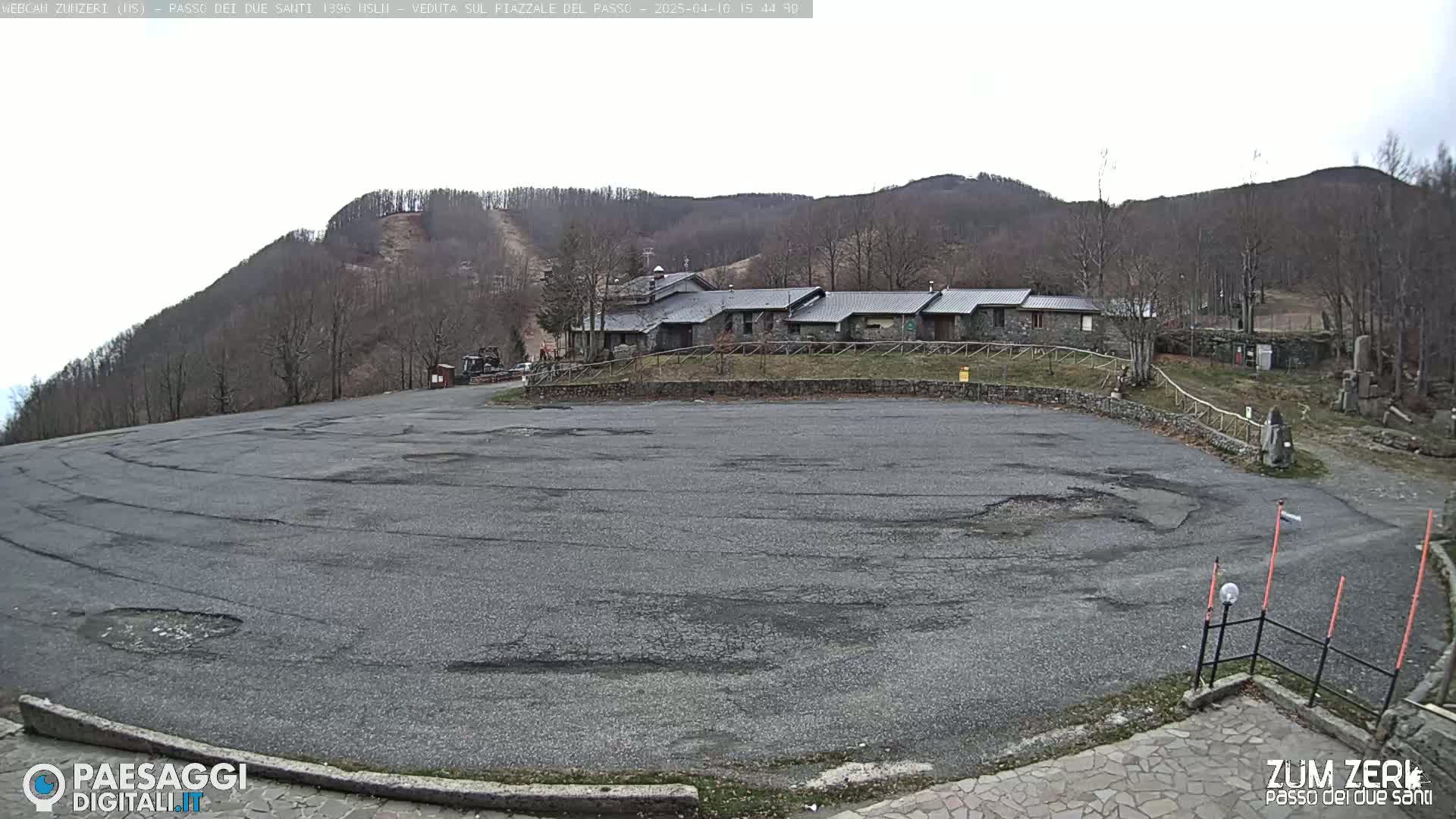 A large, empty asphalt parking lot sits before a stone building complex nestled against a tree-covered mountainside under an overcast sky.