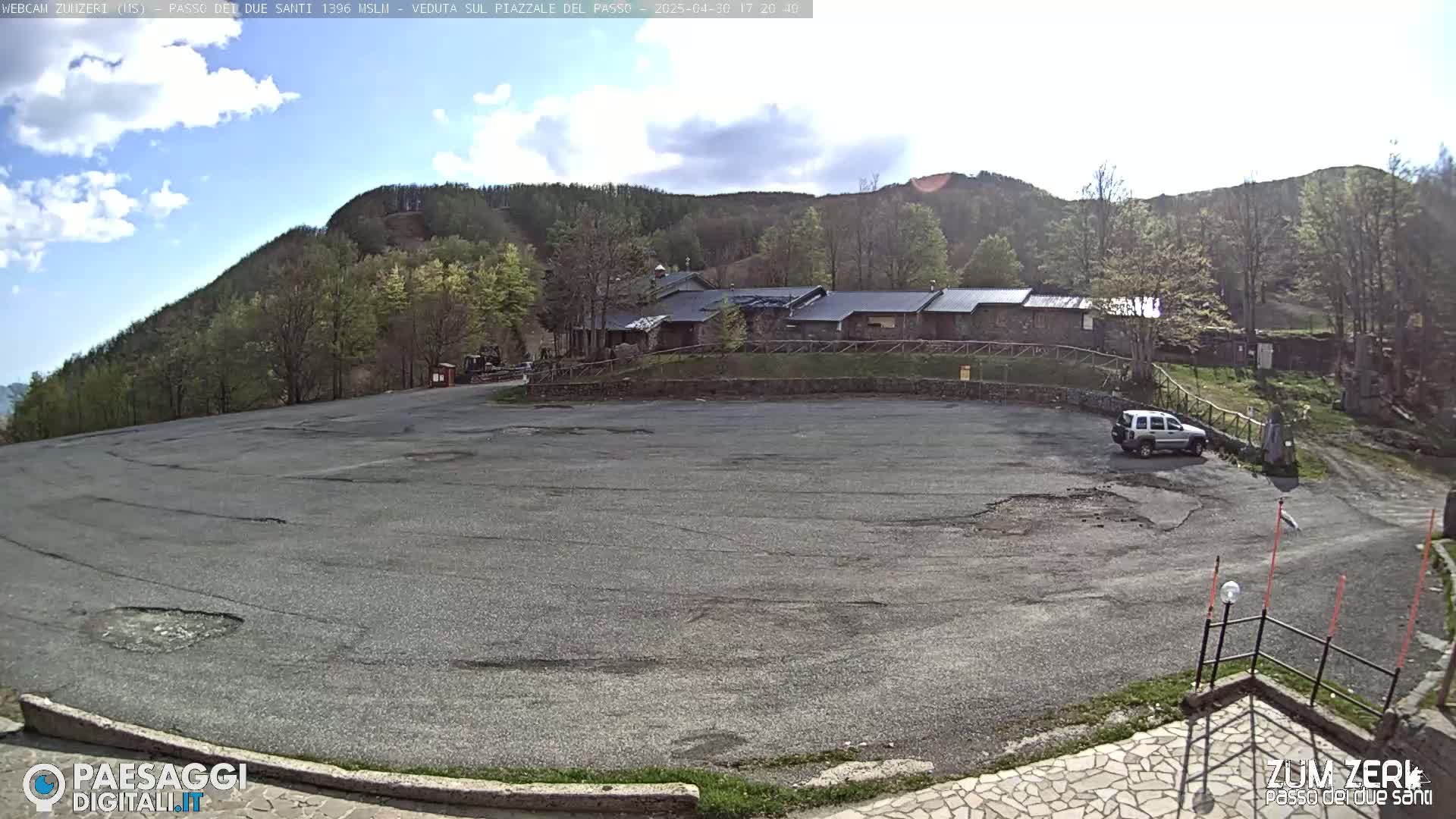 A mostly empty asphalt parking lot sits in front of a stone building complex nestled among a hillside of trees under a partly cloudy sky.