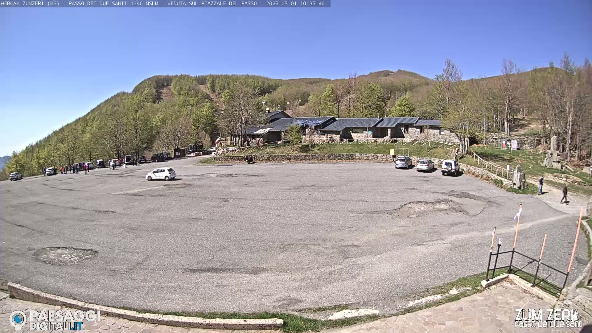 A mostly empty asphalt parking lot is surrounded by a building and a hillside covered in light green trees under a clear blue sky.