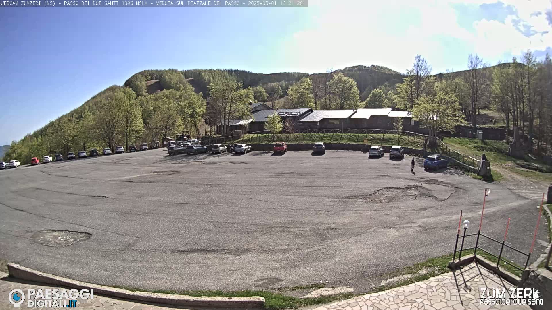 A mostly empty asphalt parking lot sits in front of a stone building nestled amongst a hillside of green trees under a sunny sky.