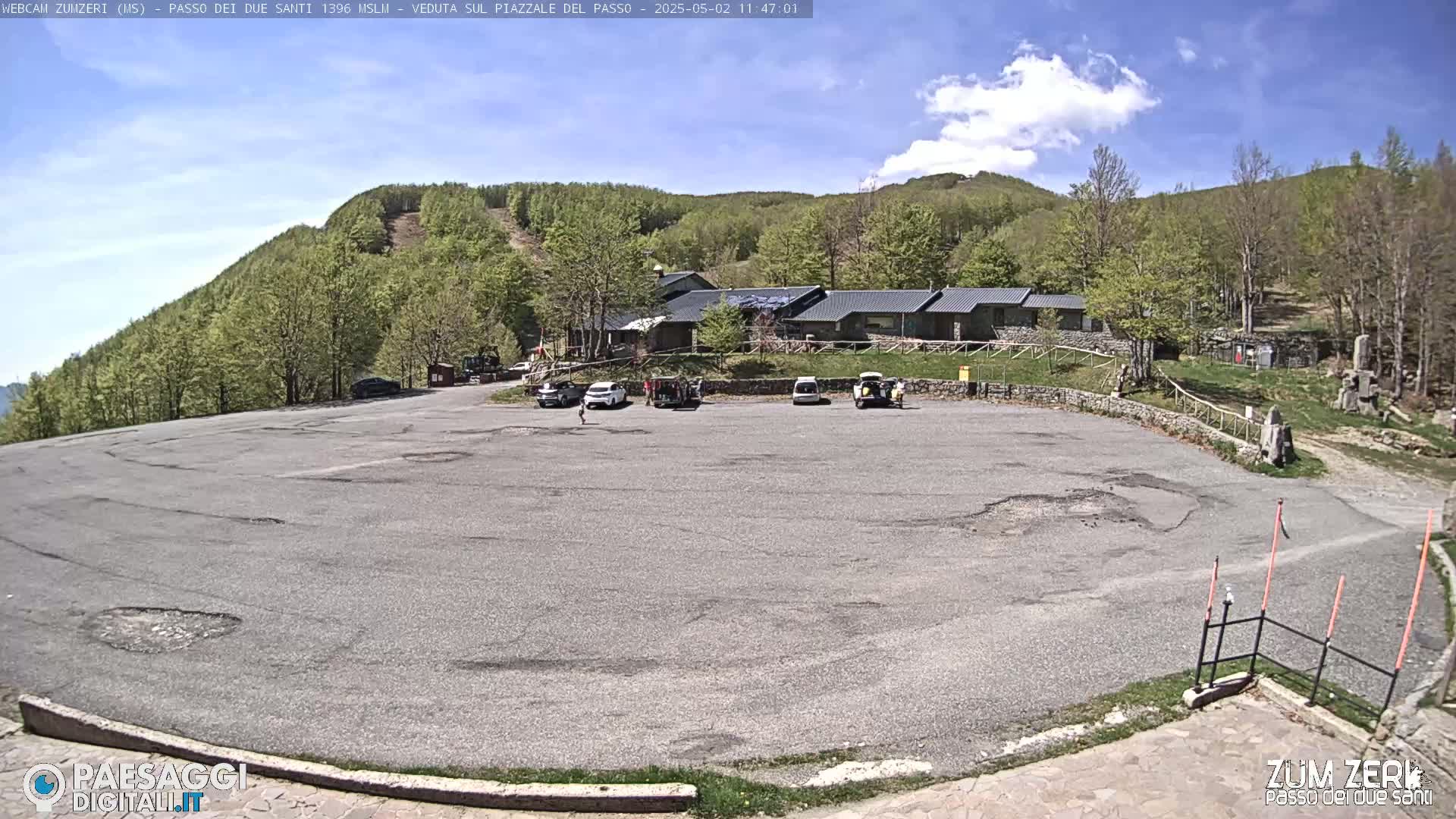 A mostly empty asphalt parking lot sits in front of a stone building, nestled amongst a verdant hillside under a bright, sunny sky.