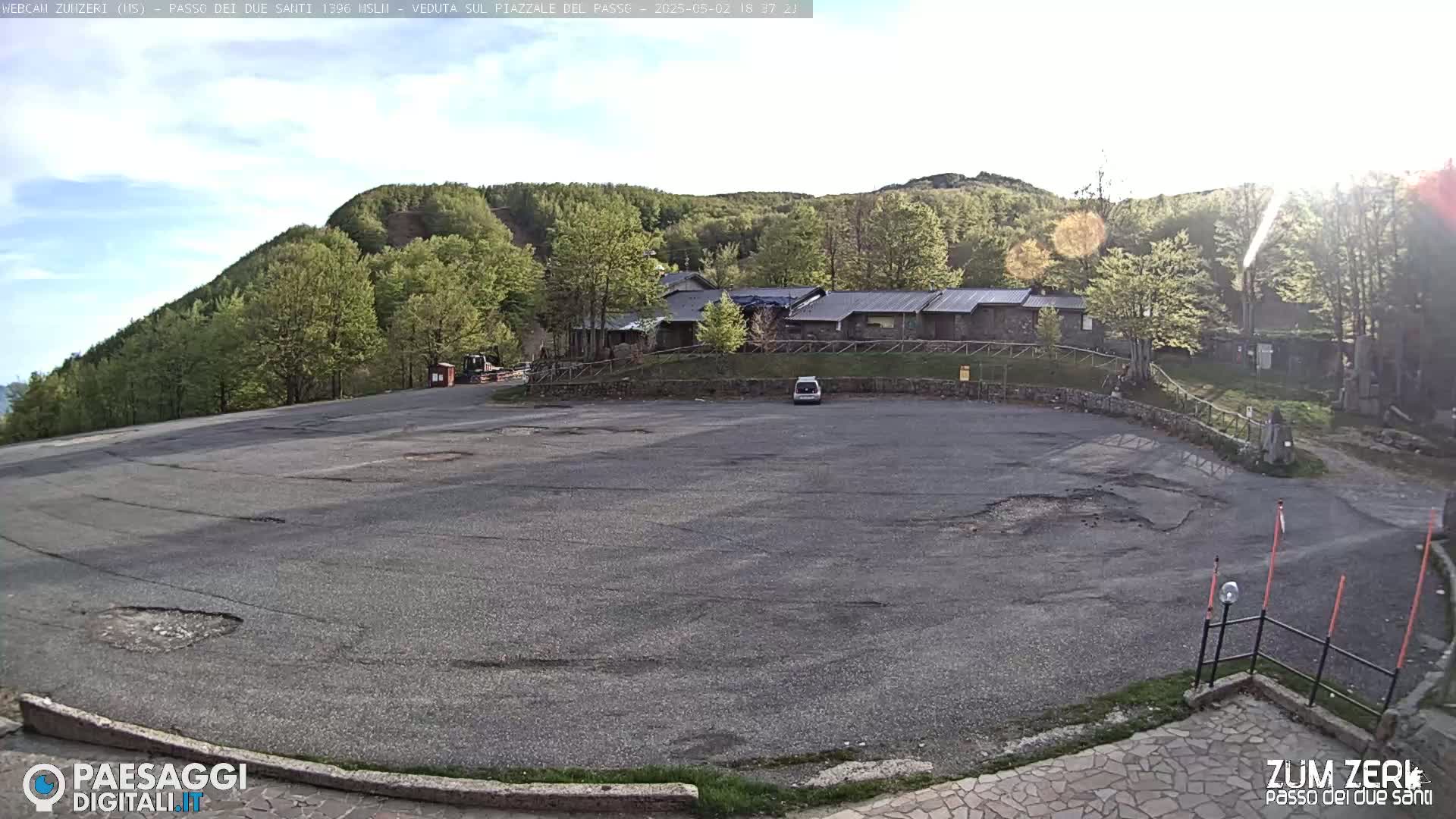 A mostly empty asphalt parking lot is situated before a building and a hillside covered in lush green trees under a partly sunny sky.