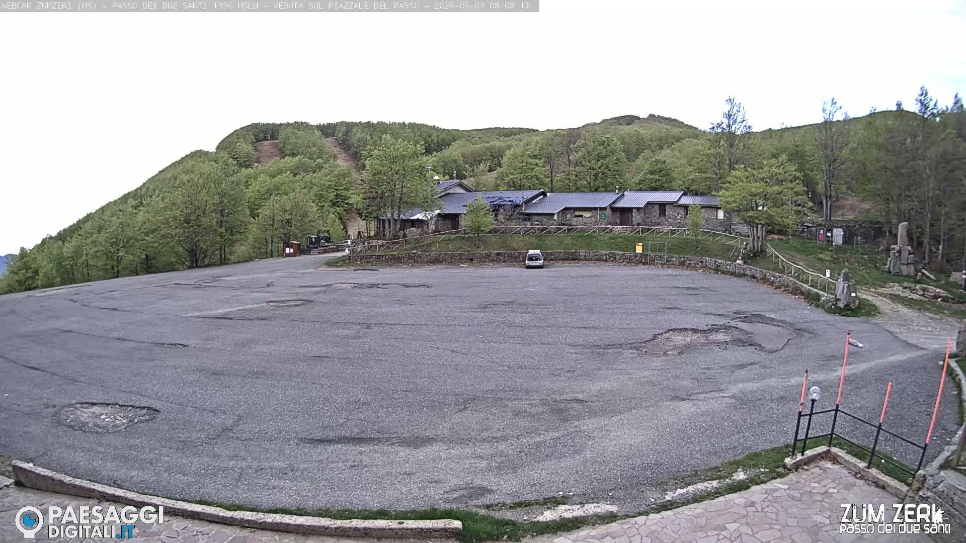 A mostly empty asphalt parking lot sits before a stone building nestled among green, leafy trees on a hill under an overcast sky.