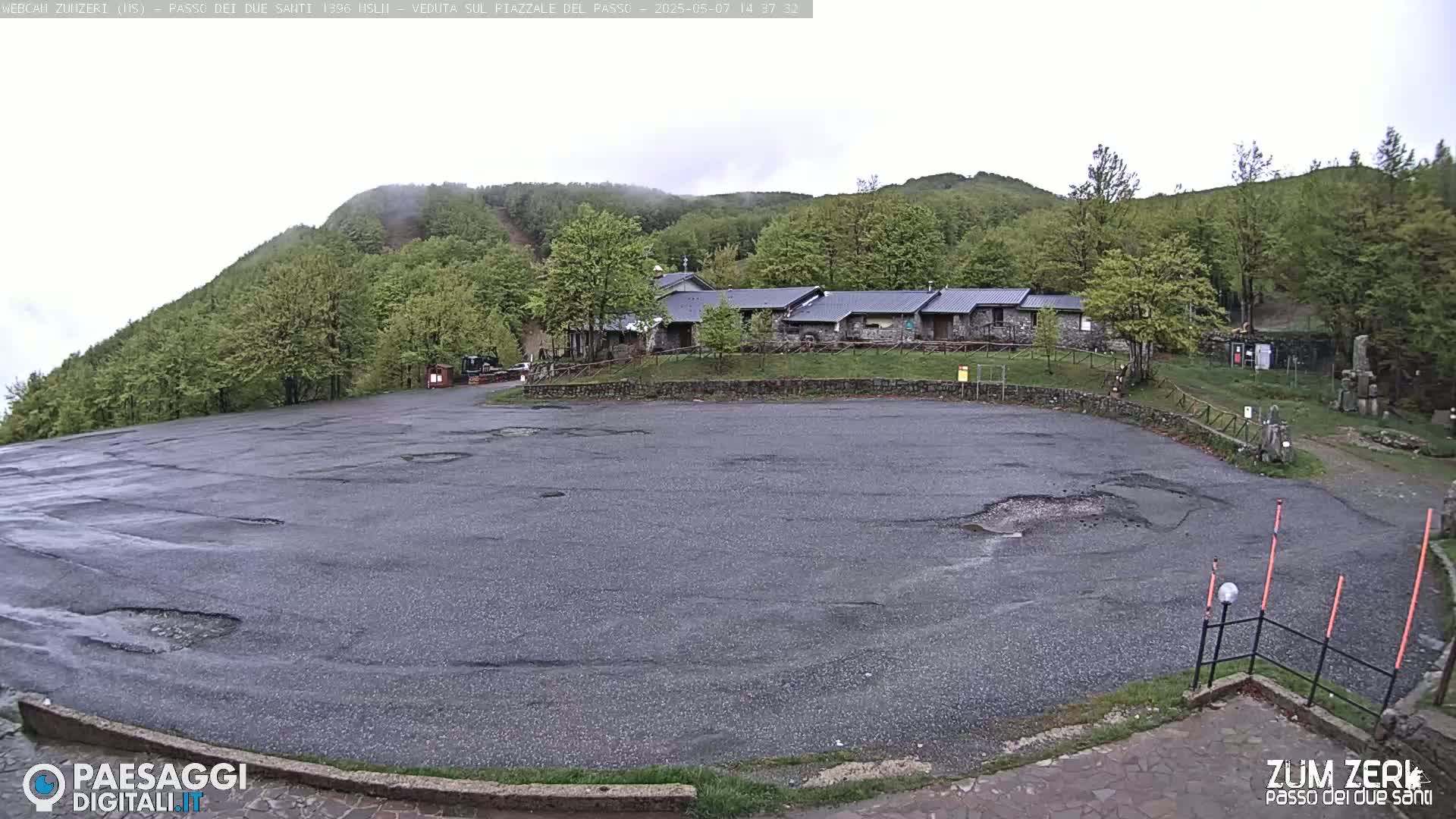A large, somewhat damaged asphalt parking lot sits before a stone building complex nestled in a green, hilly landscape under an overcast sky.