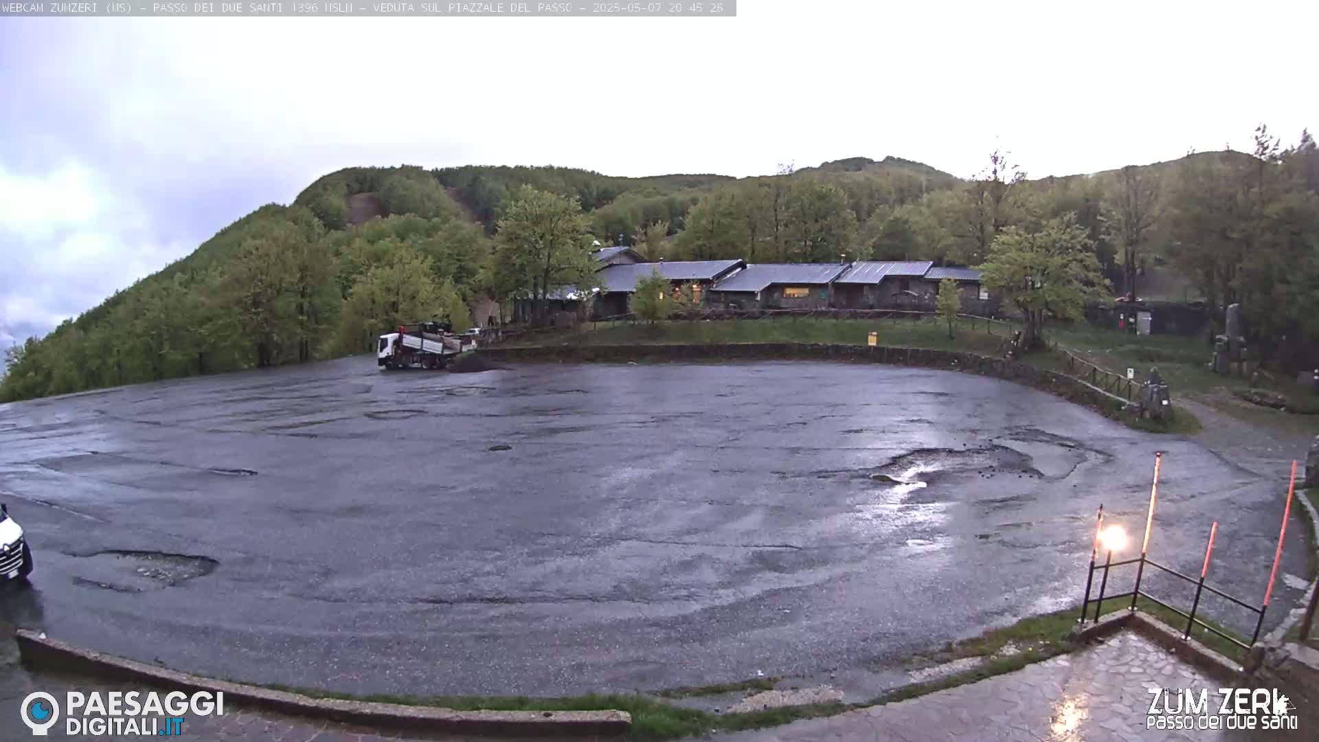 A wet, empty parking lot with potholes sits before a building complex nestled in a green, hilly landscape under an overcast sky.