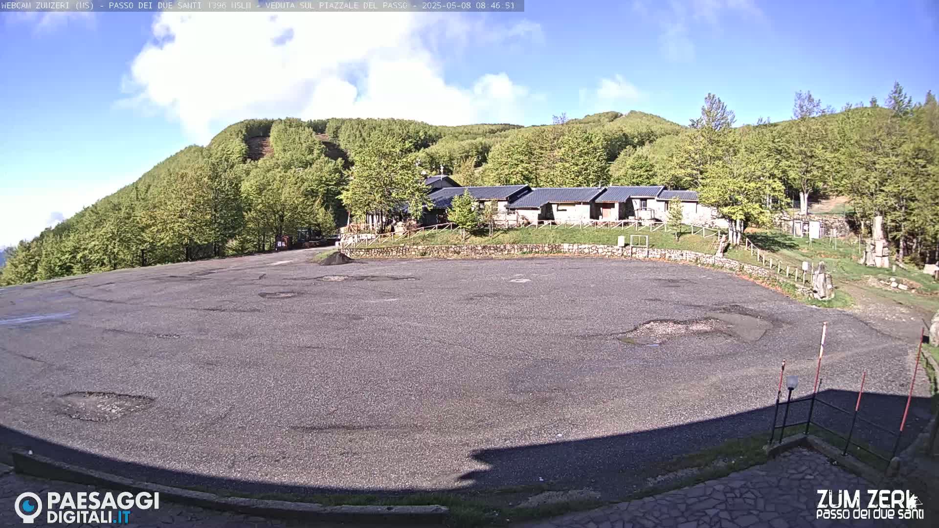 A mostly empty asphalt parking lot sits below a row of low buildings nestled in a hillside of lush green trees under a partly sunny sky.