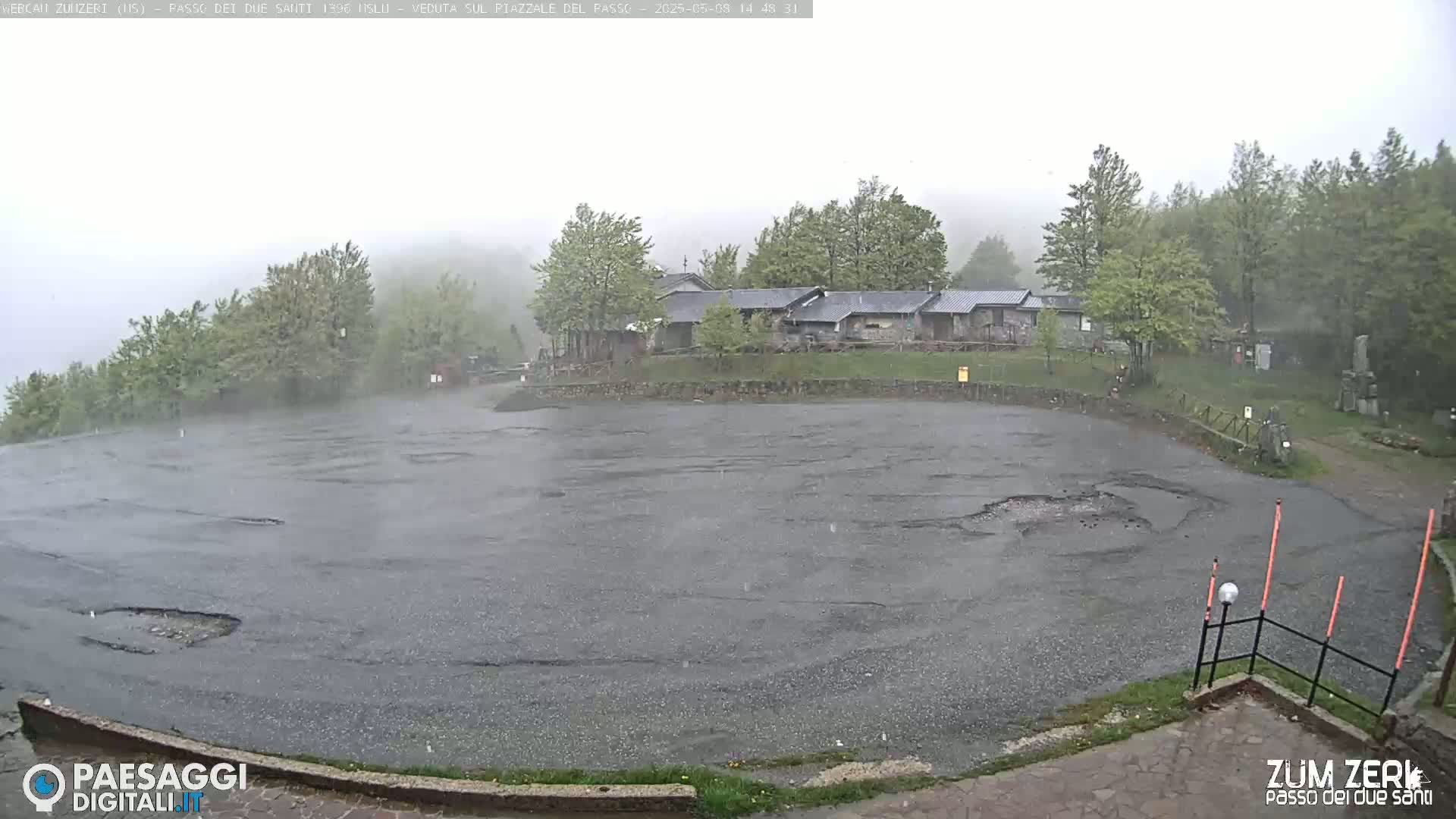 Rain falls on a large, empty asphalt lot in front of a cluster of buildings nestled among trees in a foggy mountain setting.