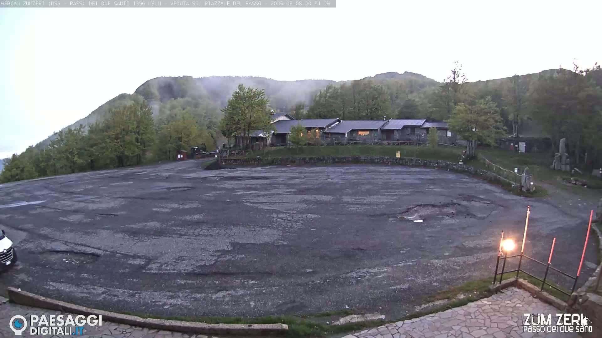 A large, cracked asphalt parking lot sits before a building complex nestled among a misty, green hillside.