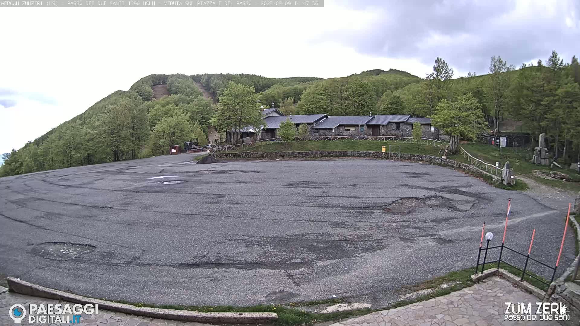 A large, cracked asphalt parking lot sits in front of a stone building complex nestled among a verdant hillside under an overcast sky.