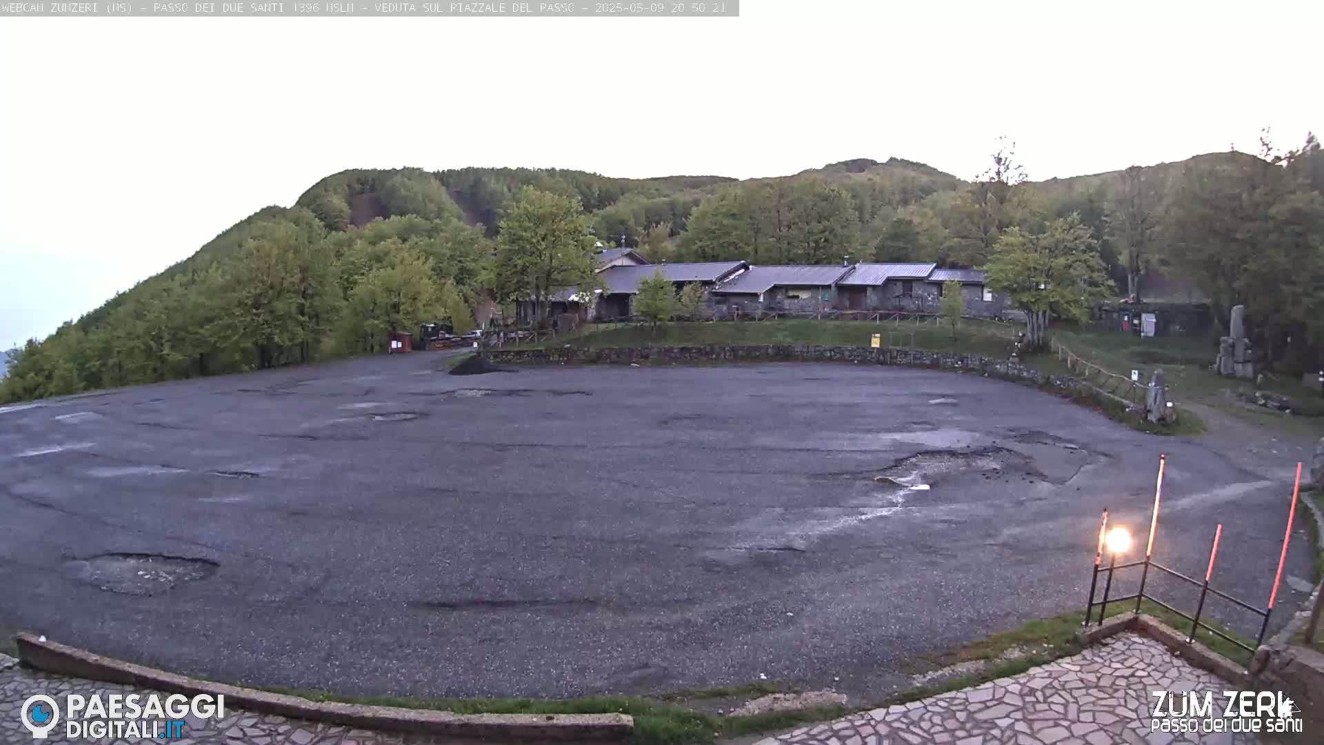 A large, potholed asphalt parking lot sits before a cluster of low stone buildings nestled against a verdant hillside under an overcast sky.