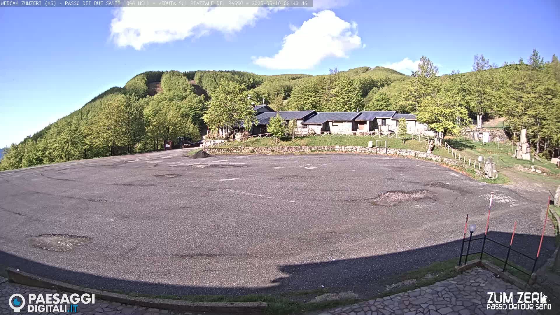 A large, empty asphalt parking lot is situated in front of a cluster of buildings, all surrounded by a lush green forest under a mostly sunny sky.