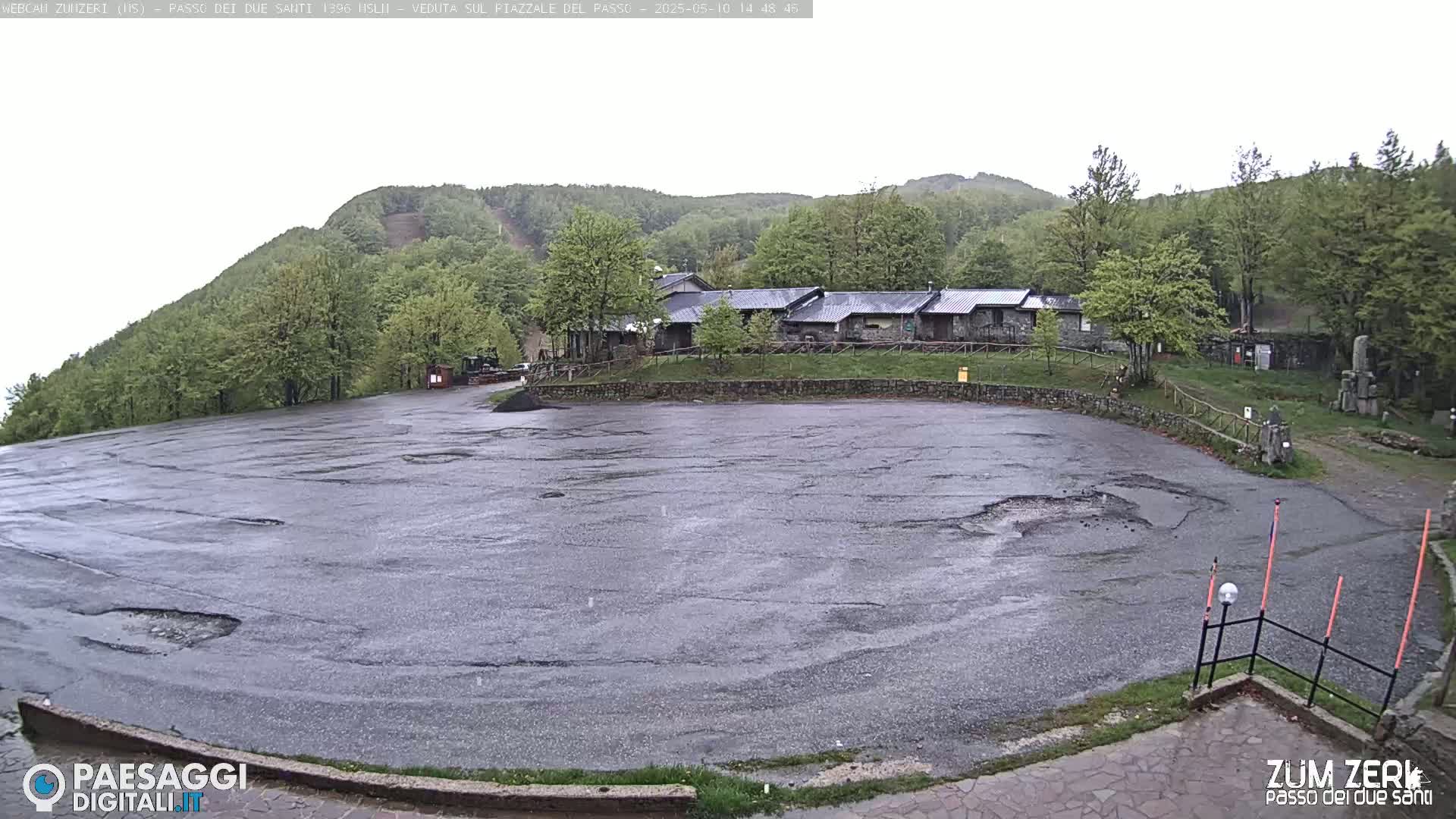 A wet, empty asphalt parking lot sits before a cluster of buildings nestled among green hills under an overcast sky.