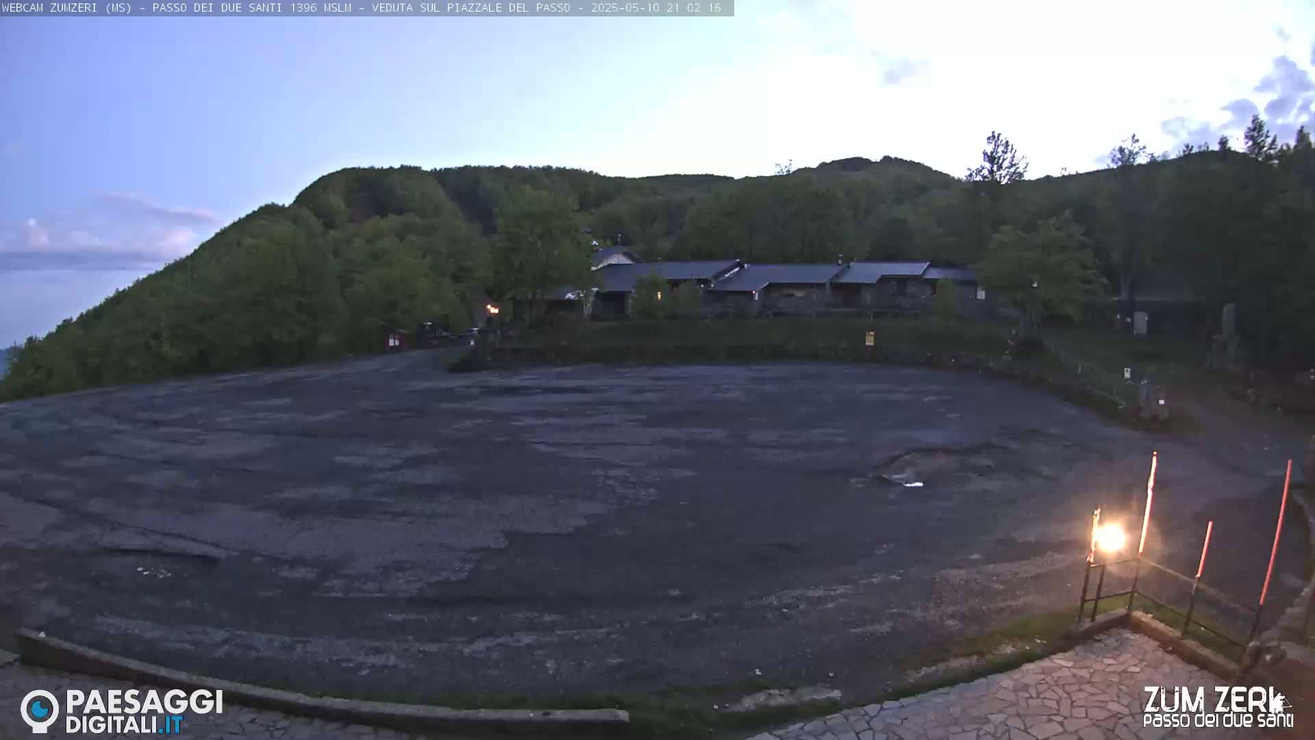 A large, empty asphalt lot sits before a stone building nestled in a green, hilly landscape under a twilight sky.