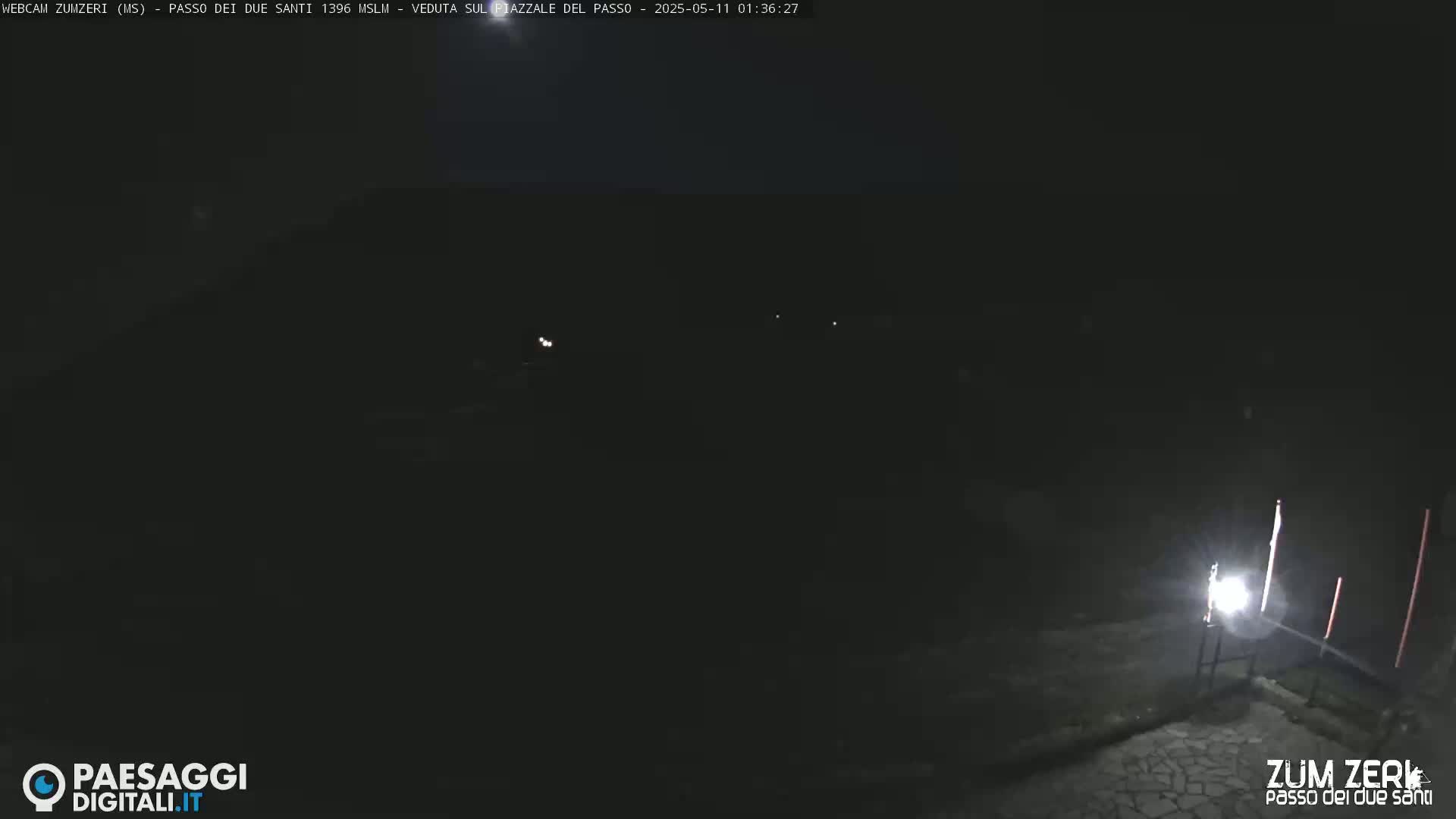 A dark, nighttime scene shows a dimly lit area with a few faint lights visible in the distance.