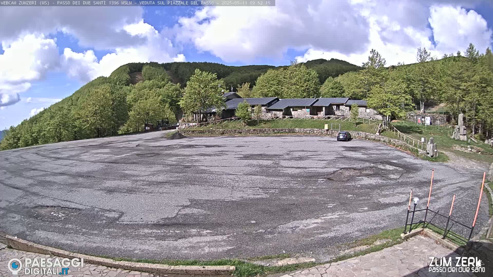 A large, empty asphalt parking lot sits before a low stone building complex nestled in a verdant, tree-covered hillside under a partly cloudy sky.