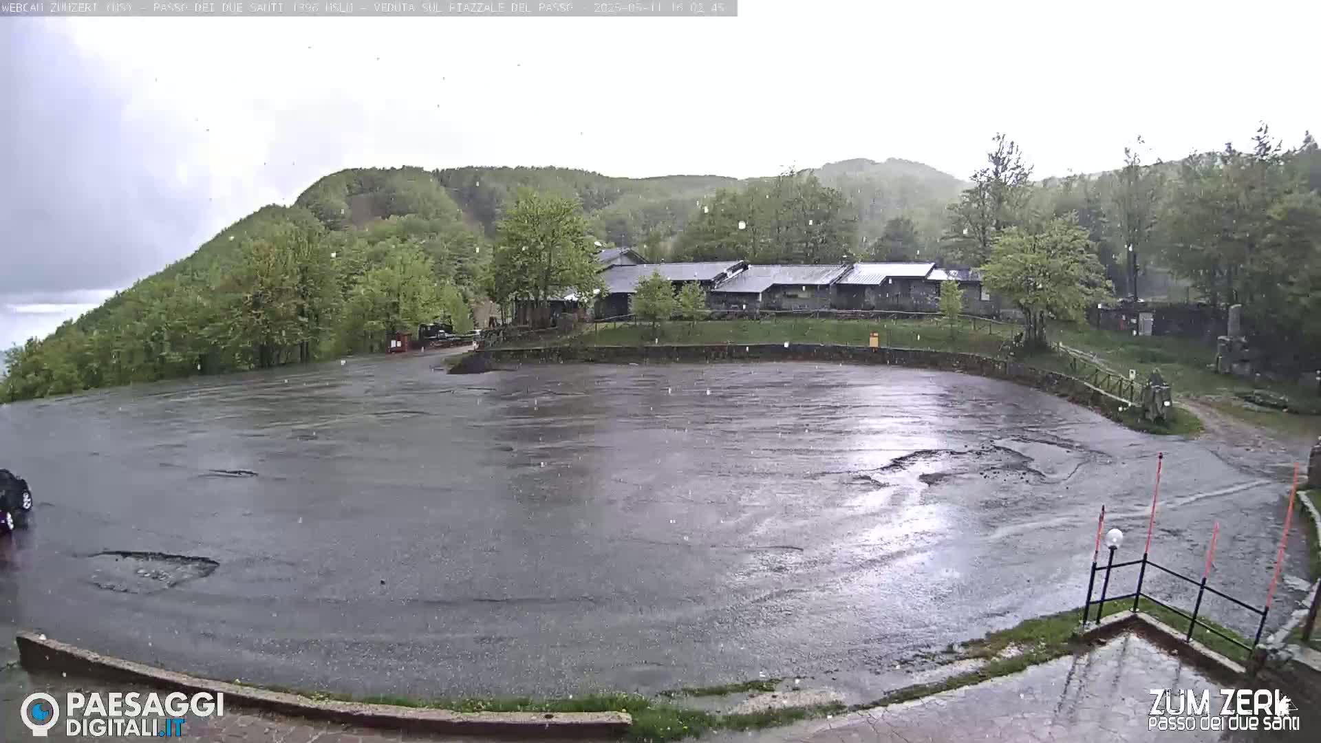 A wet, empty asphalt parking lot is seen during a light rain shower, with a building and a wooded hillside in the background.