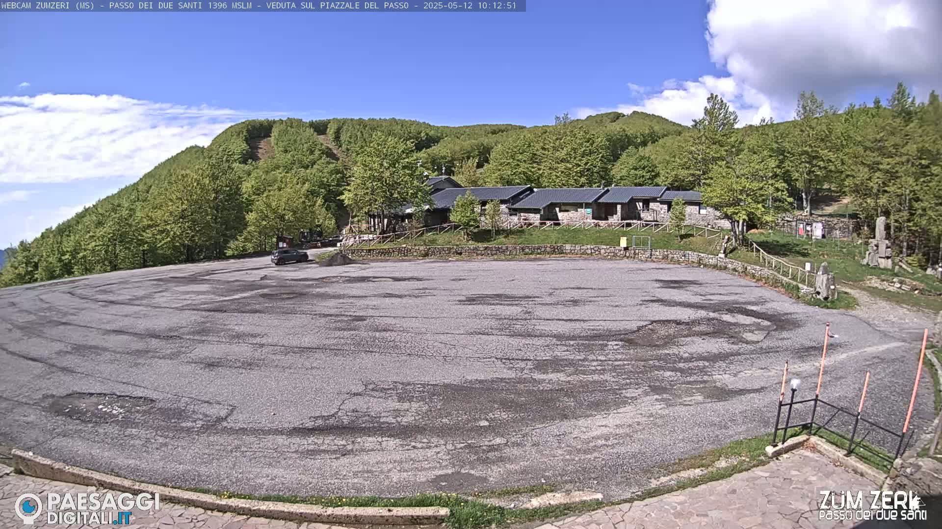 A mostly empty paved parking lot sits before a cluster of low buildings nestled amongst a verdant, tree-covered hillside under a mostly sunny sky.