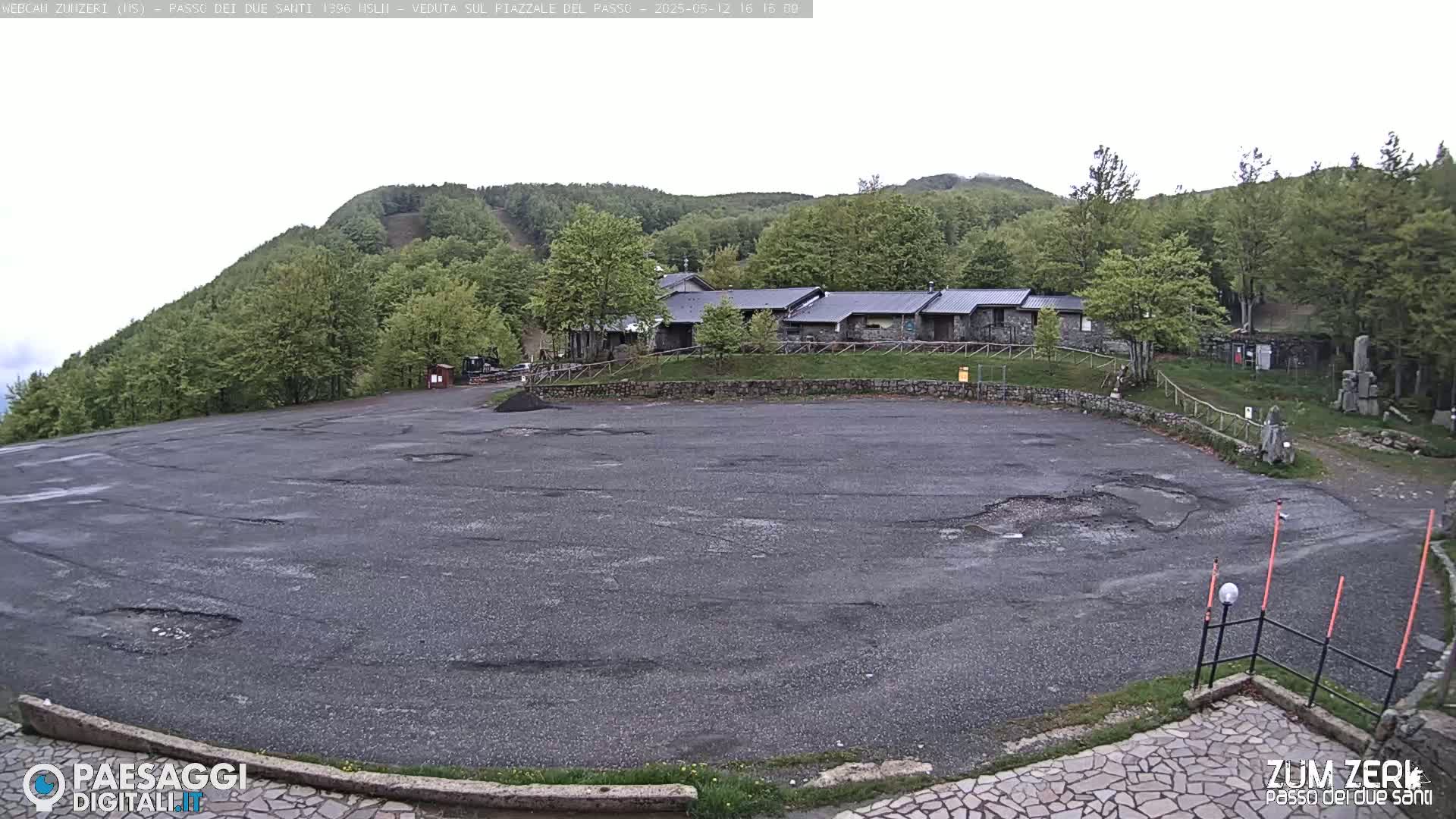 A large, empty asphalt parking lot sits before a row of buildings nestled in a green, hilly forest under an overcast sky.