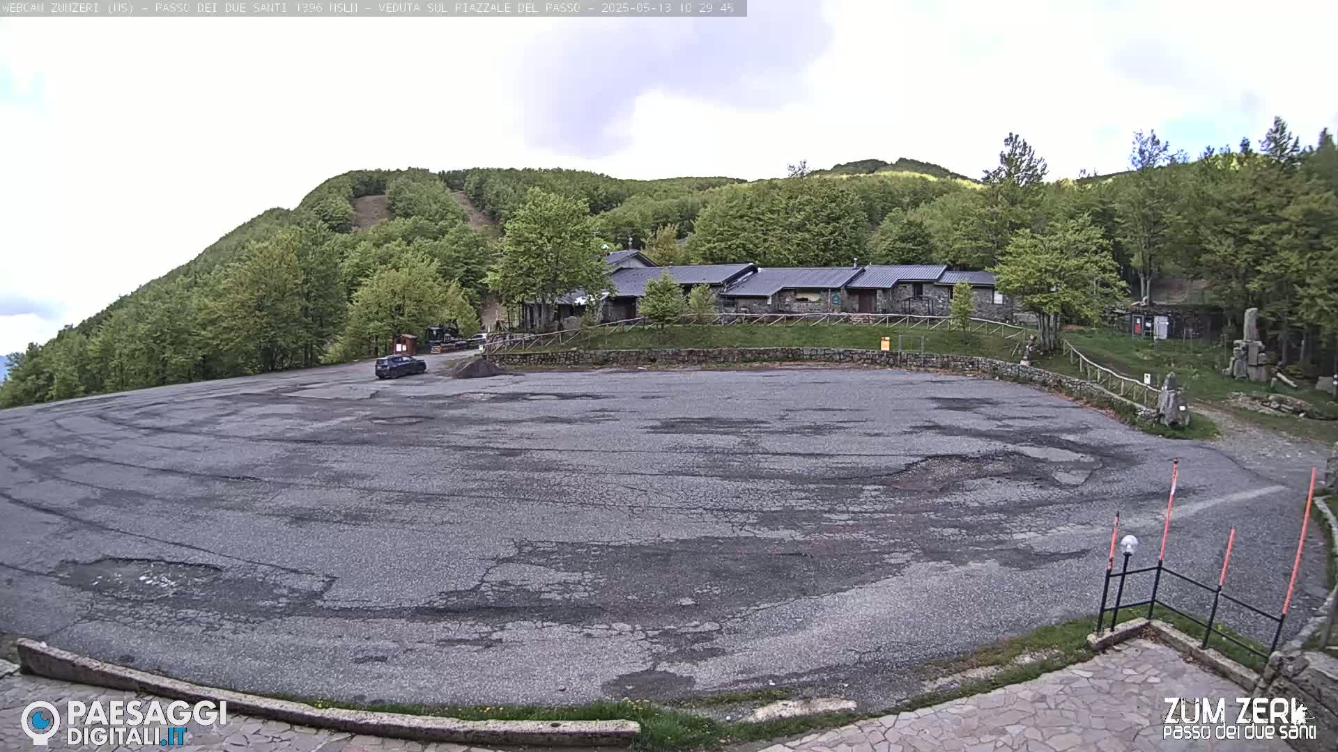 A large, cracked asphalt parking lot sits before a stone building complex nestled in a hillside of lush green trees under an overcast sky.