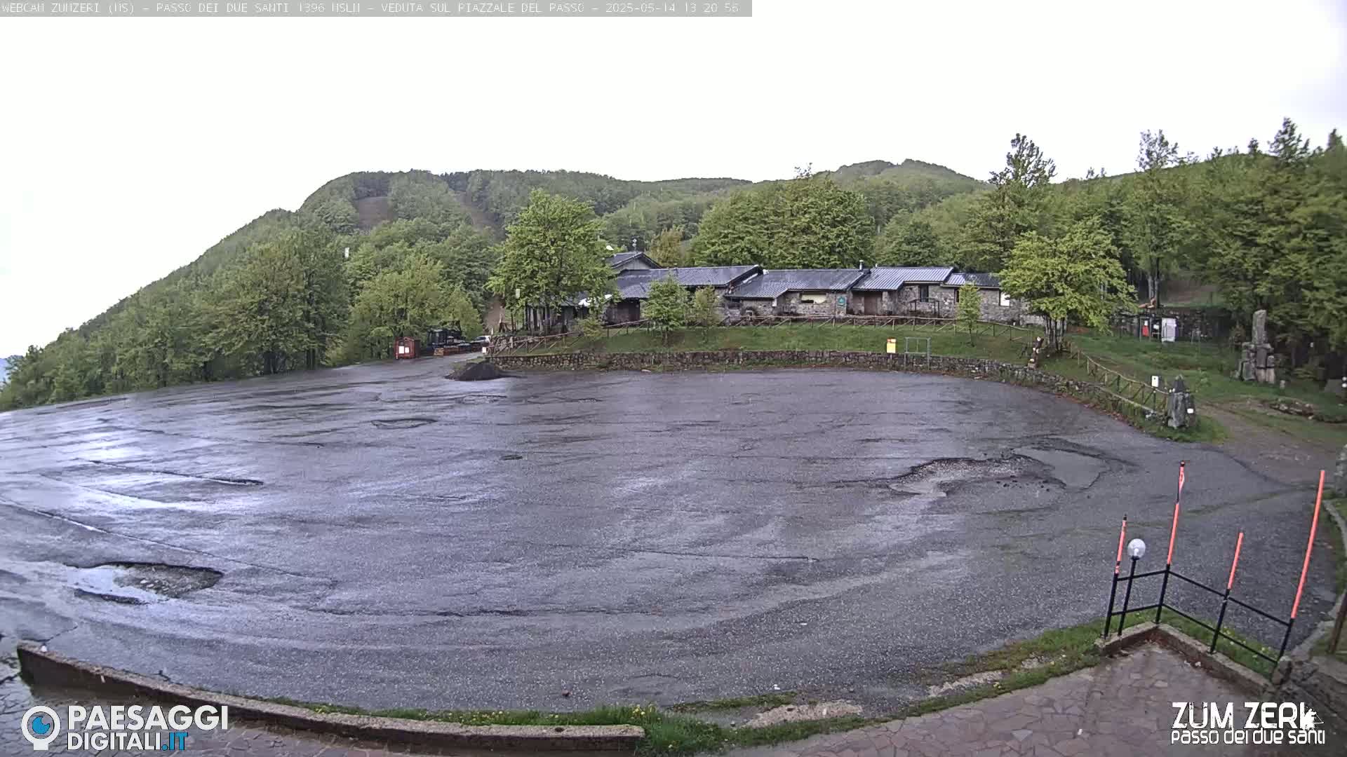 A wet, empty asphalt parking lot sits before a stone building nestled amongst lush green trees and hills under an overcast sky.