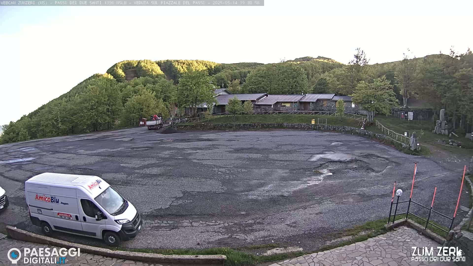 A mostly empty asphalt parking lot sits below a building nestled amongst green hills and trees under a mostly clear sky.