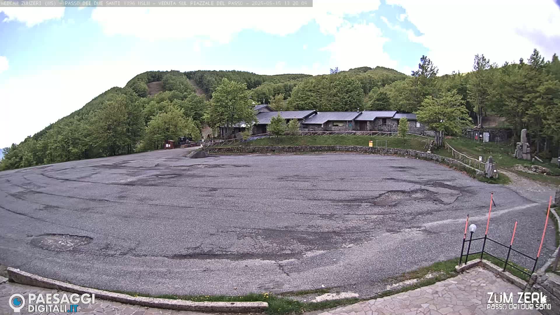 A large, cracked asphalt parking lot sits before a low building complex nestled amongst a verdant hillside under a partly cloudy sky.