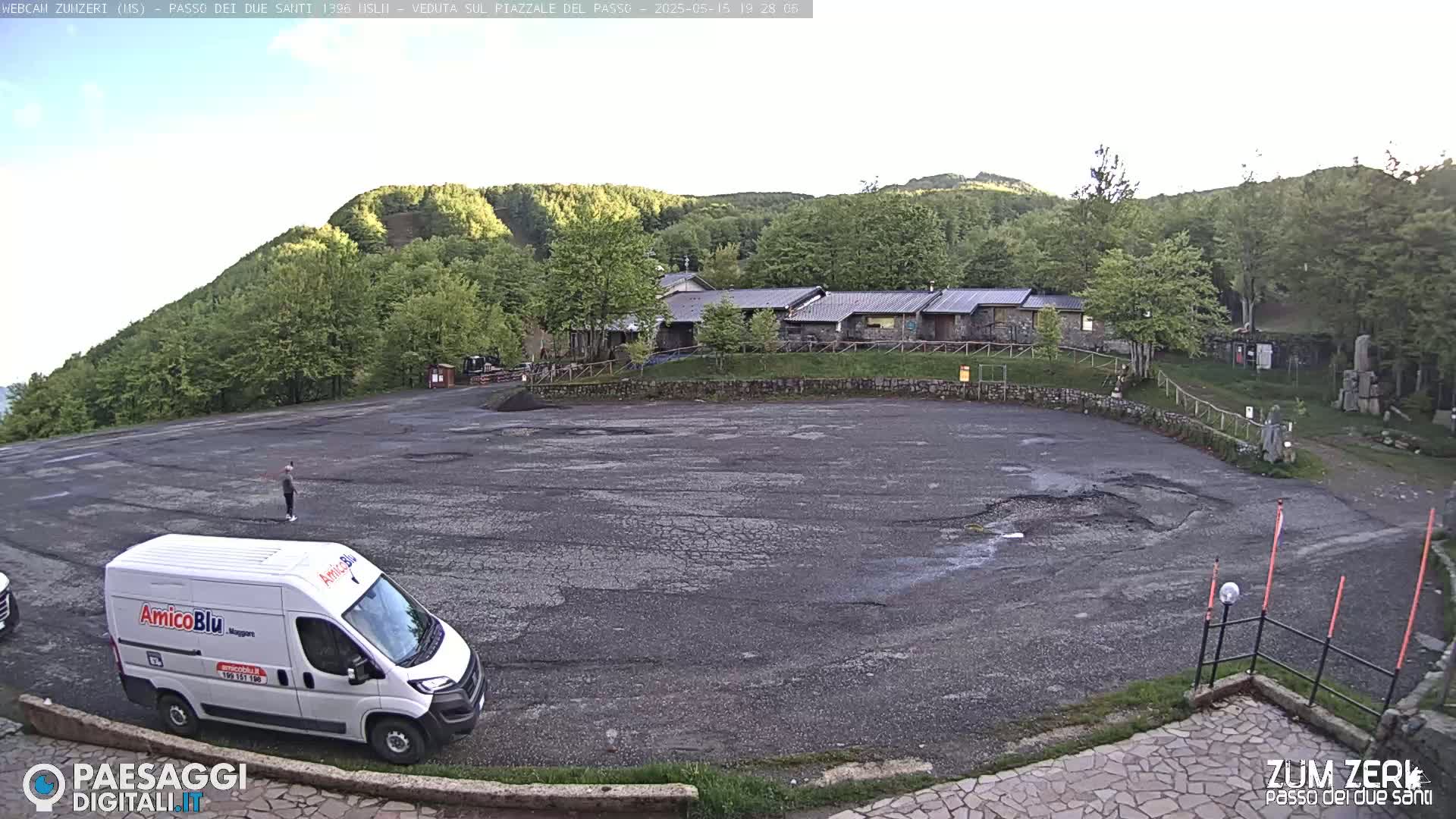 A large, empty asphalt parking lot sits below a building nestled among a lush green forest under a mostly clear sky.