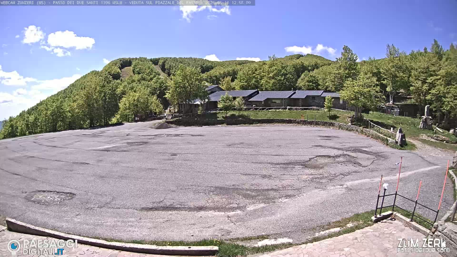 A large, empty asphalt parking lot sits below a cluster of buildings nestled amongst a verdant, hilly forest under a partly cloudy sky.