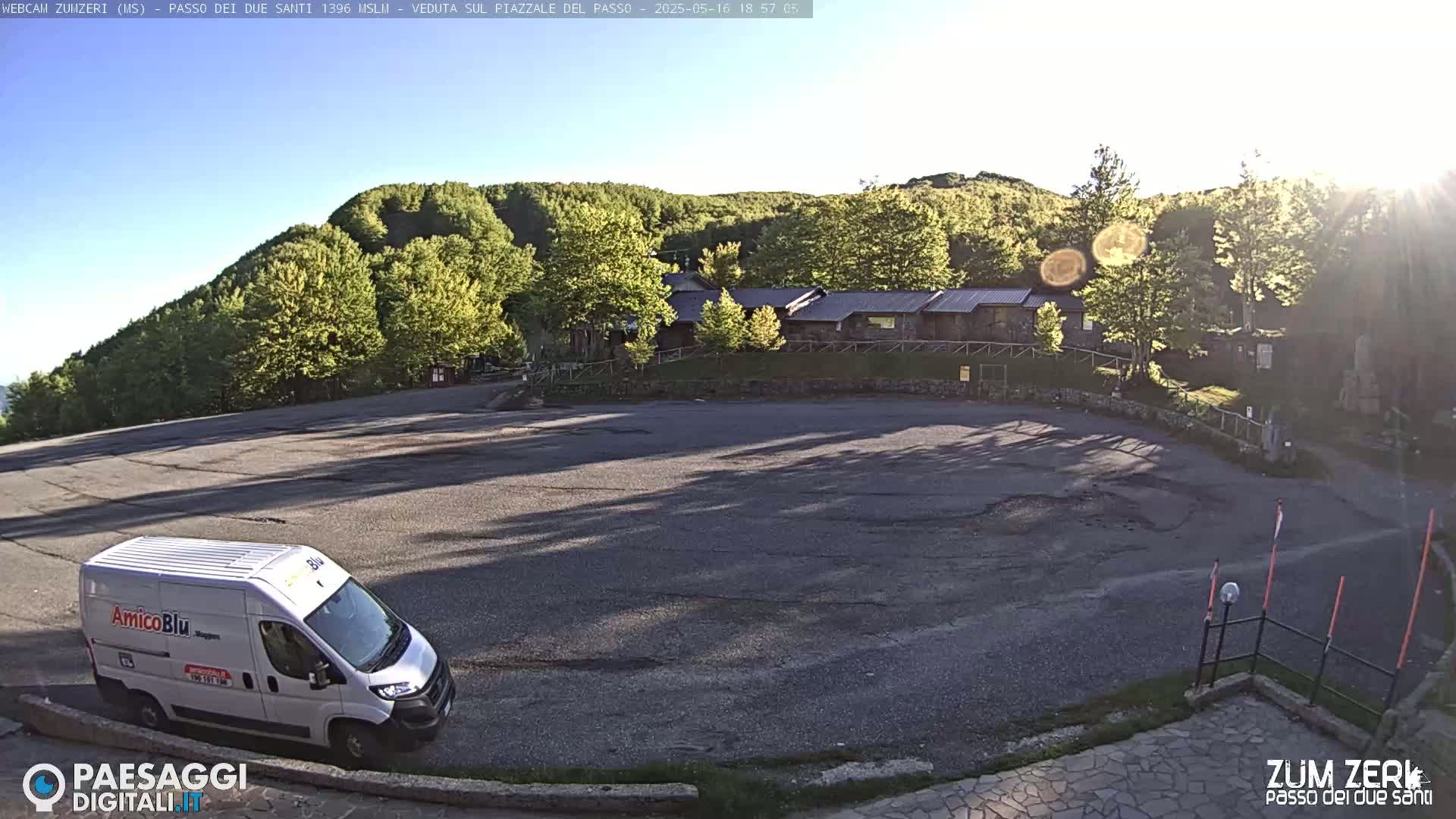 A white van is parked in a mostly empty asphalt lot near a building complex nestled among green hills under a clear, sunny sky.