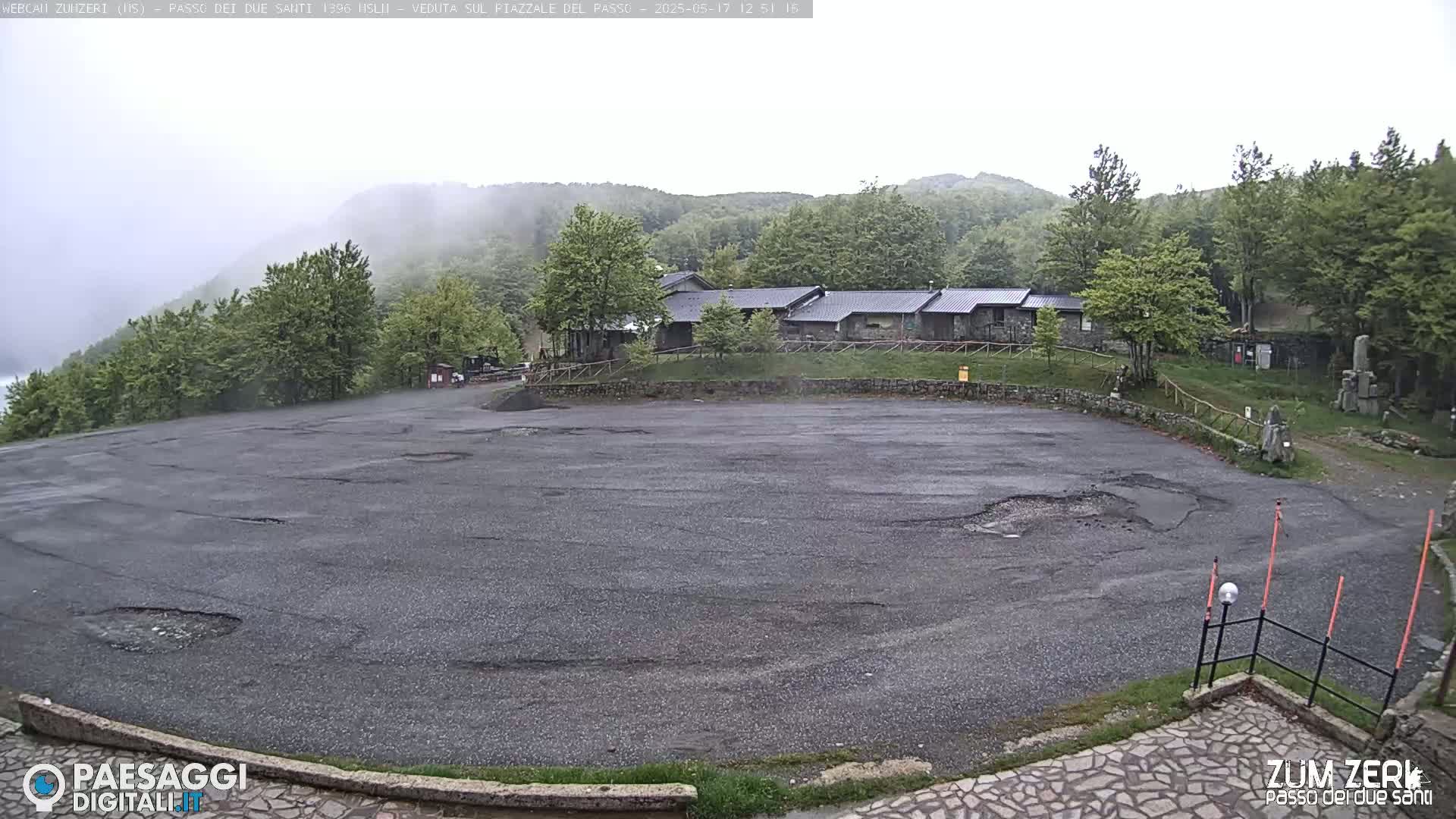 A foggy, overcast day reveals a large, damaged asphalt parking lot overlooking a building complex nestled among green, wooded hills.