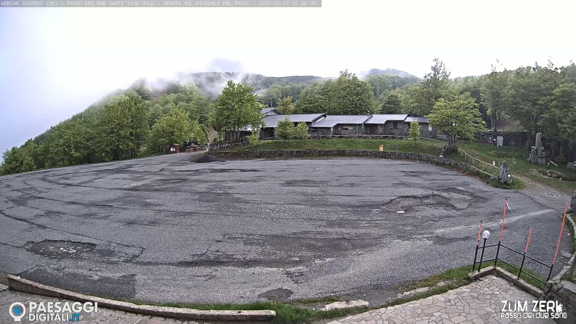 A mostly empty, cracked asphalt parking lot sits before a row of stone buildings nestled among a green, fog-shrouded hillside.