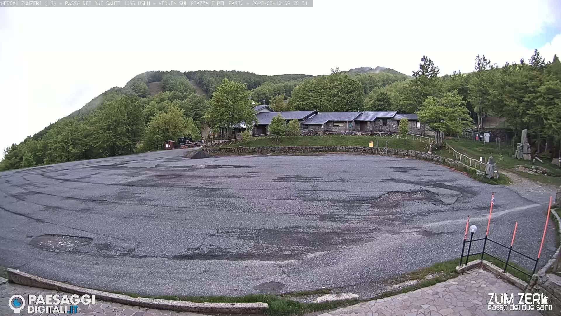 An empty asphalt parking lot with some cracks sits before a stone building complex nestled among green, leafy trees on a slightly overcast day.